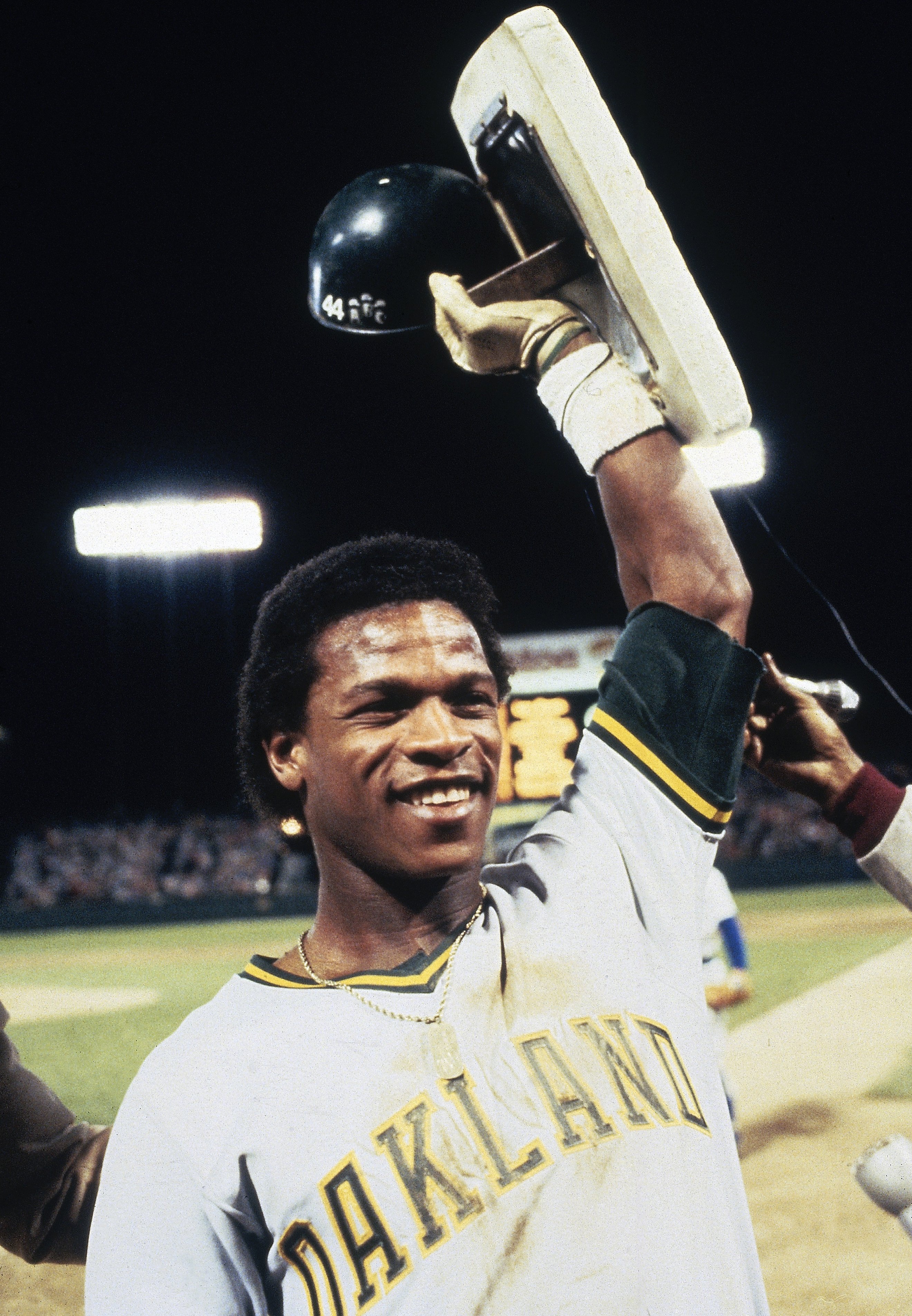 A baseball player in an Oakland jersey smiles and raises a base and helmet above his head. The stadium lights shine brightly in the background.