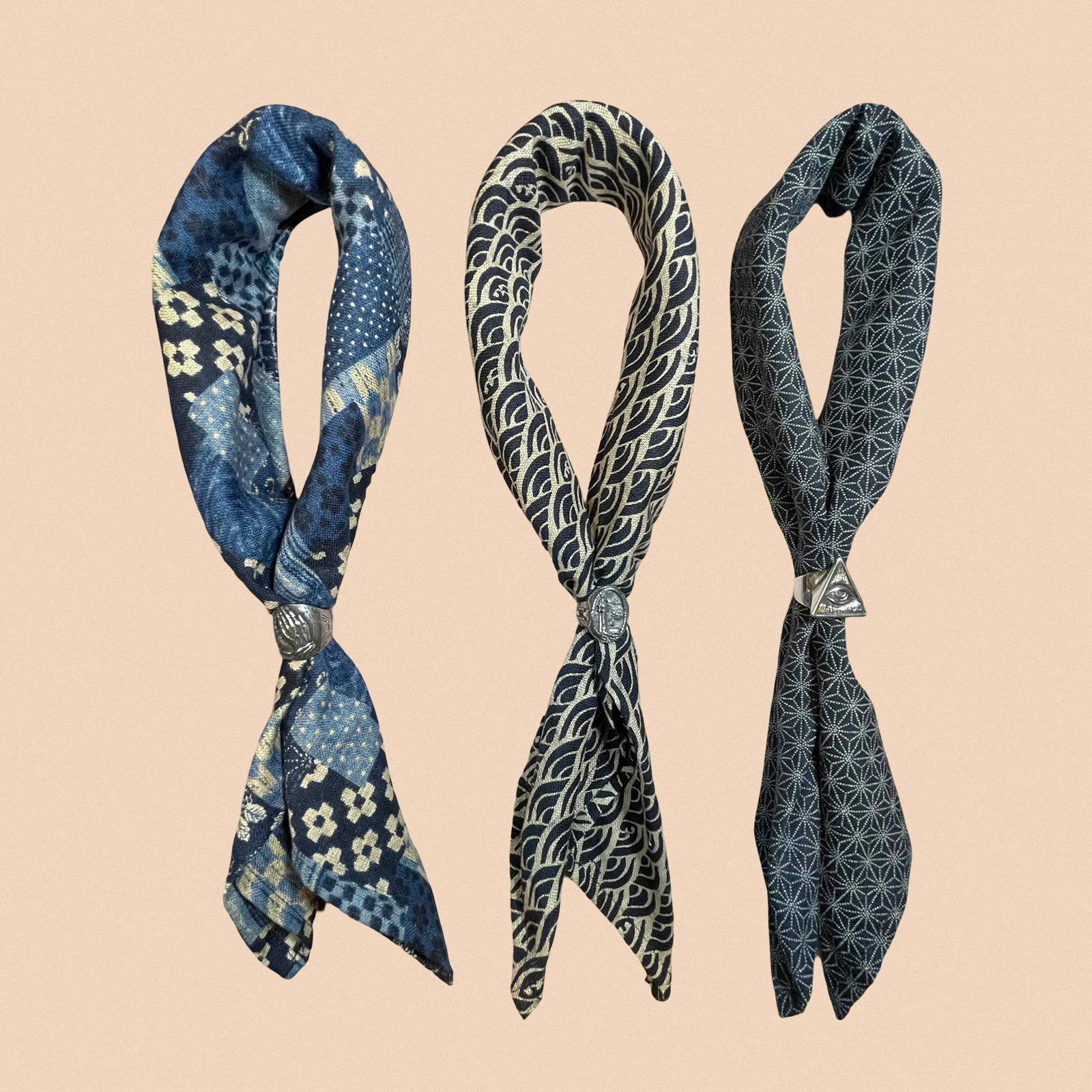 Three patterned scarves hang vertically with metal rings. The left scarf is blue with floral designs, the center has wave patterns, and the right features geometric shapes.