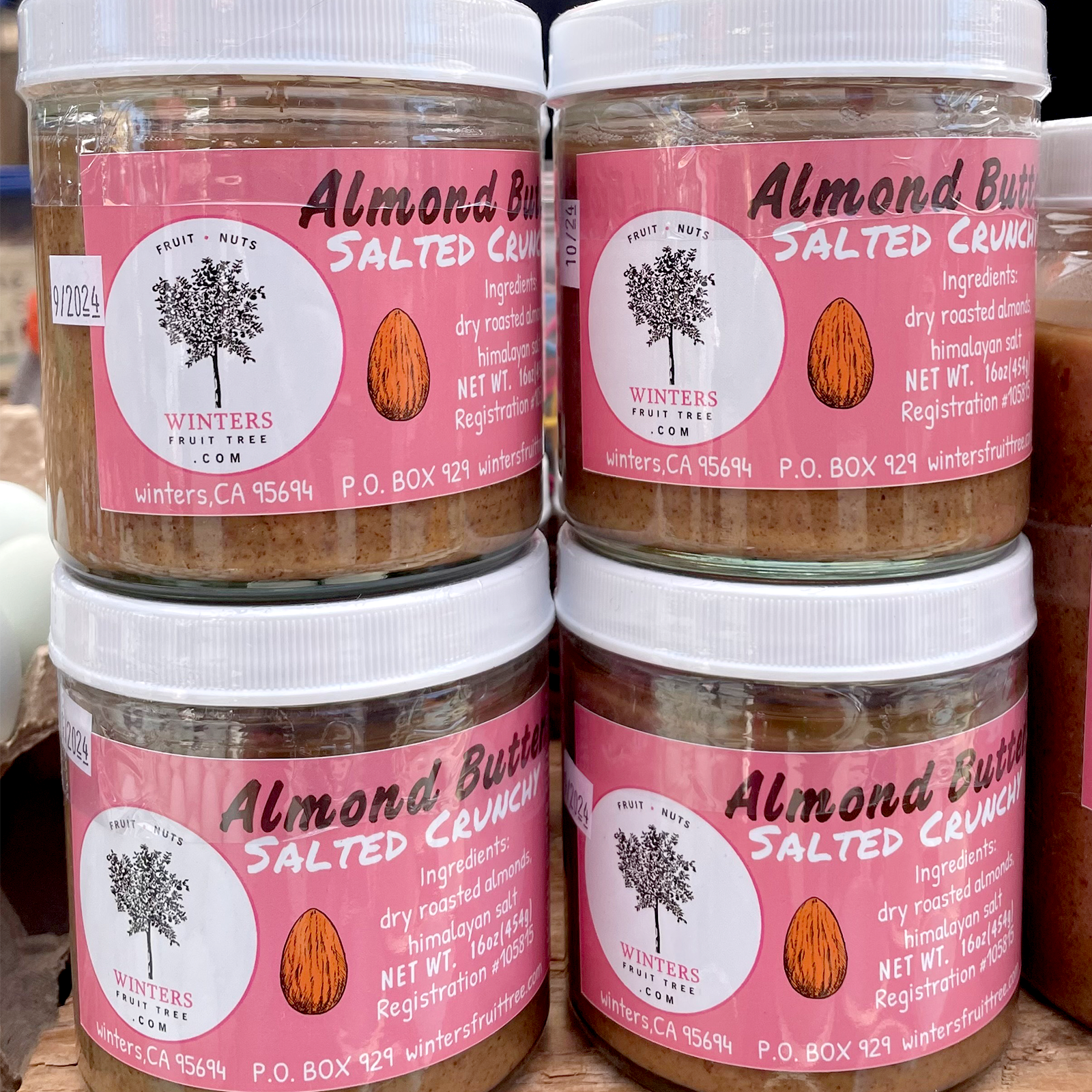 This image shows jars of almond butter labeled &quot;Salted Crunchy Almond Butter&quot; by Winters Fruit Tree. The labels are pink with white lids, featuring an almond and tree logo.