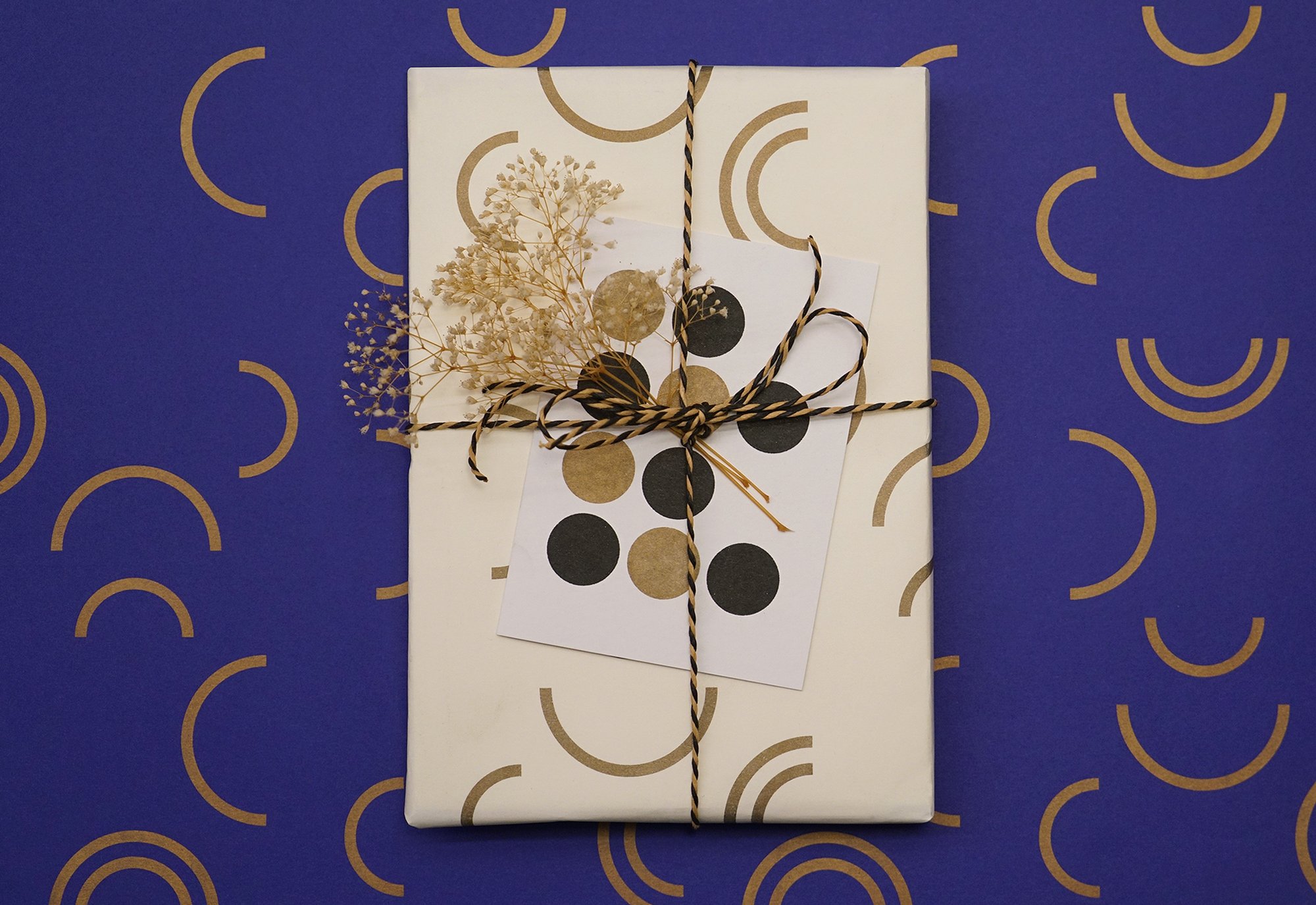 A gift wrapped in white paper with brown semi-circle patterns sits on a blue background with yellow semi-circles. It is tied with twine, adorned with dried flowers and a polka dot card.