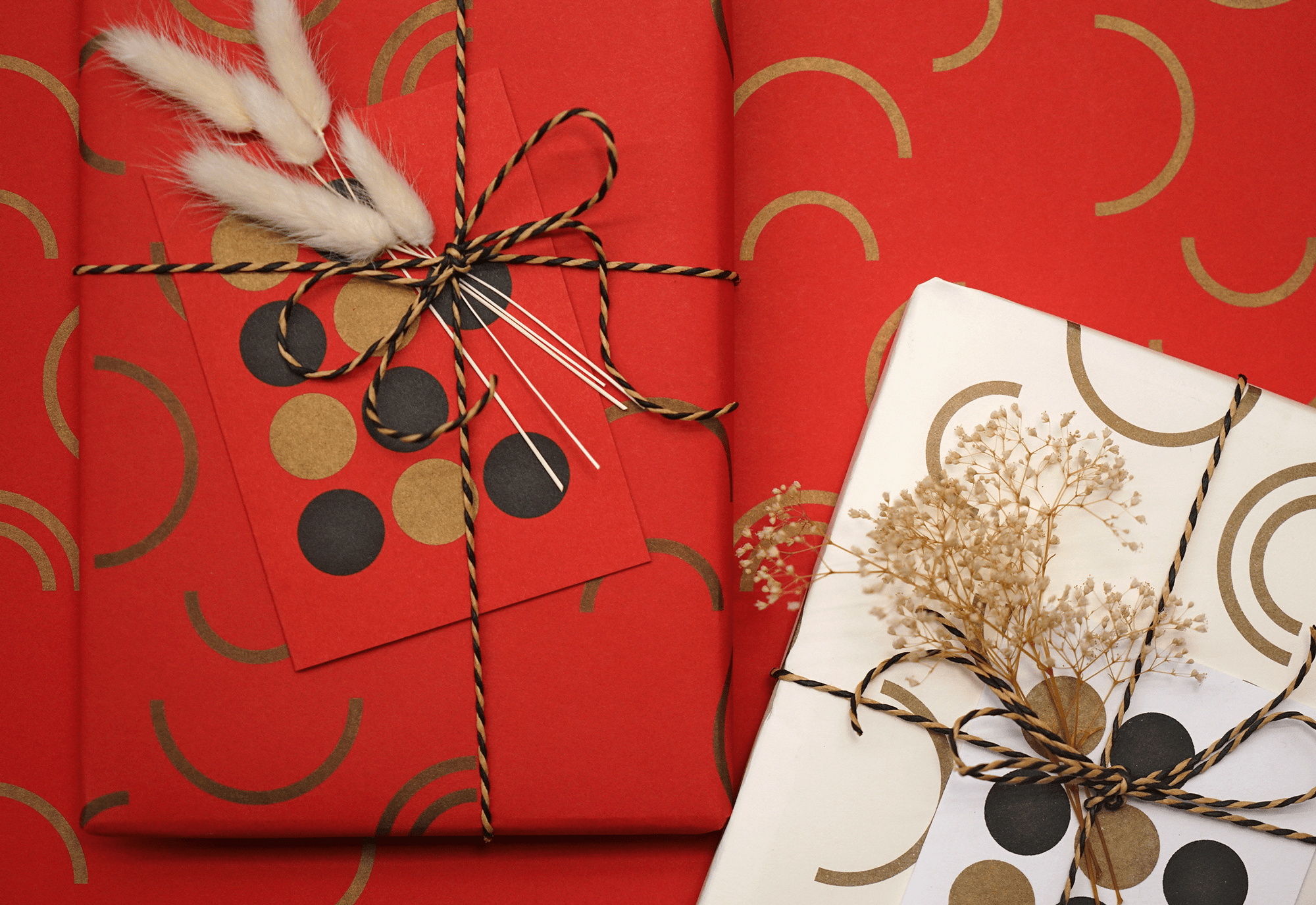 A gift wrapped in red paper with gold and black arc patterns, tied with a black-and-gold twine bow, decorated with white feathers and a card with dots.