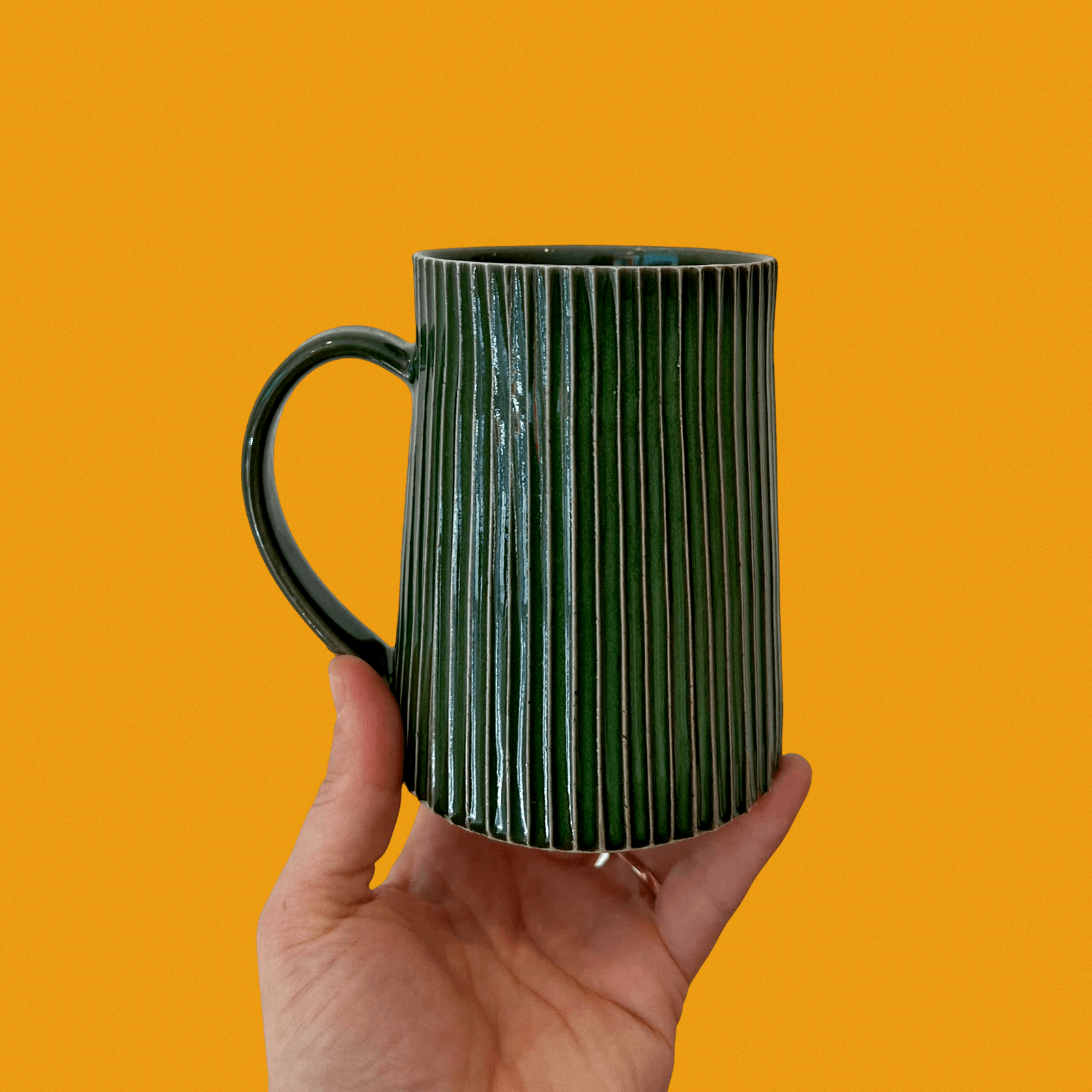 A person is holding a dark green, vertically ribbed ceramic mug against a bright orange background. The mug has a large handle and a glossy finish.