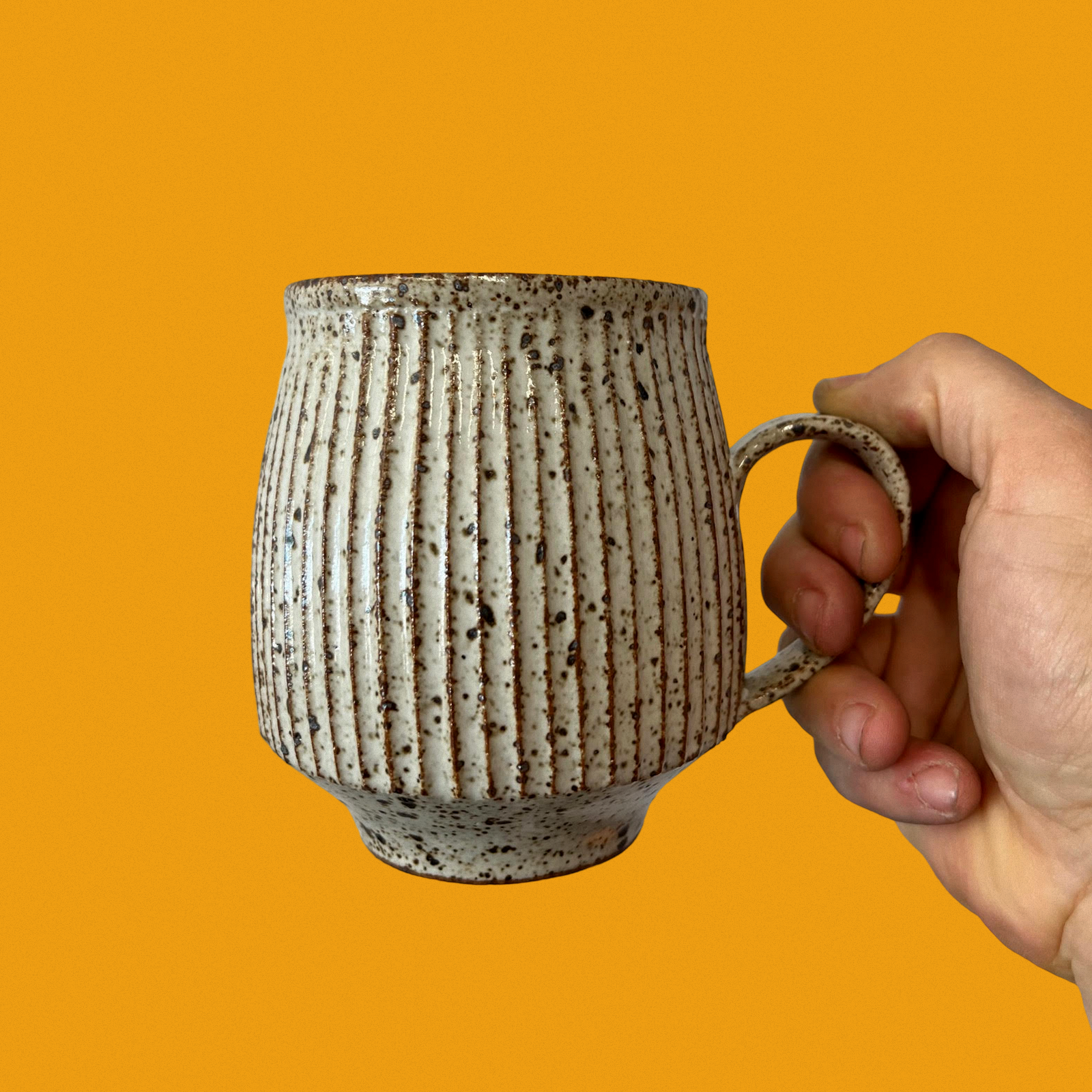 A hand holds a speckled ceramic mug with vertical ridges against a bright orange background.