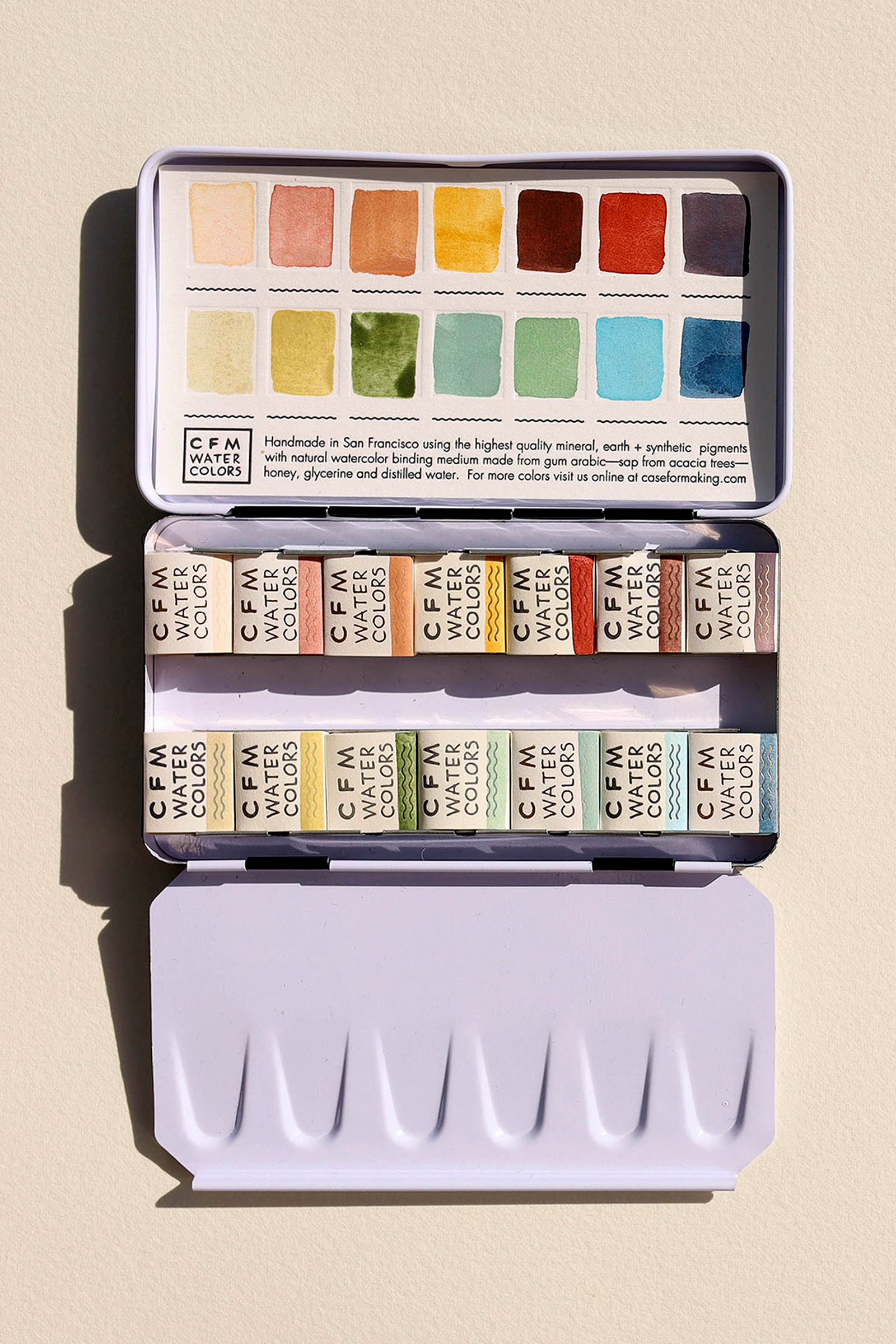 The image shows an open tin containing a watercolor palette with organized color pans in shades like browns, greens, and blues. The lid displays color samples.