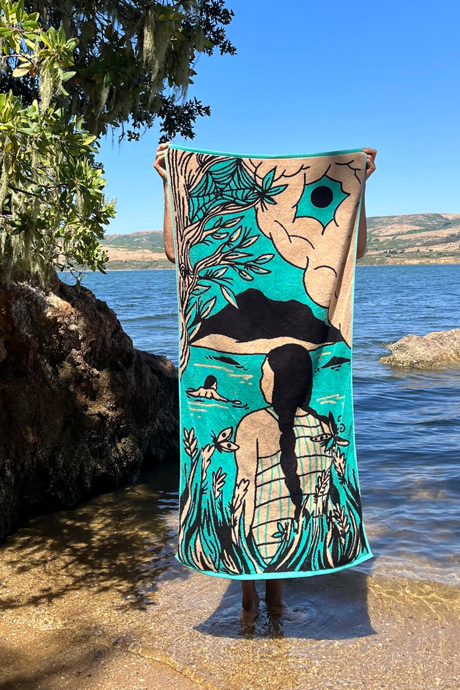 A person stands in shallow water, holding a colorful towel with a nature scene of a figure gazing at mountains and sky. Tree branches frame the lake in the background.