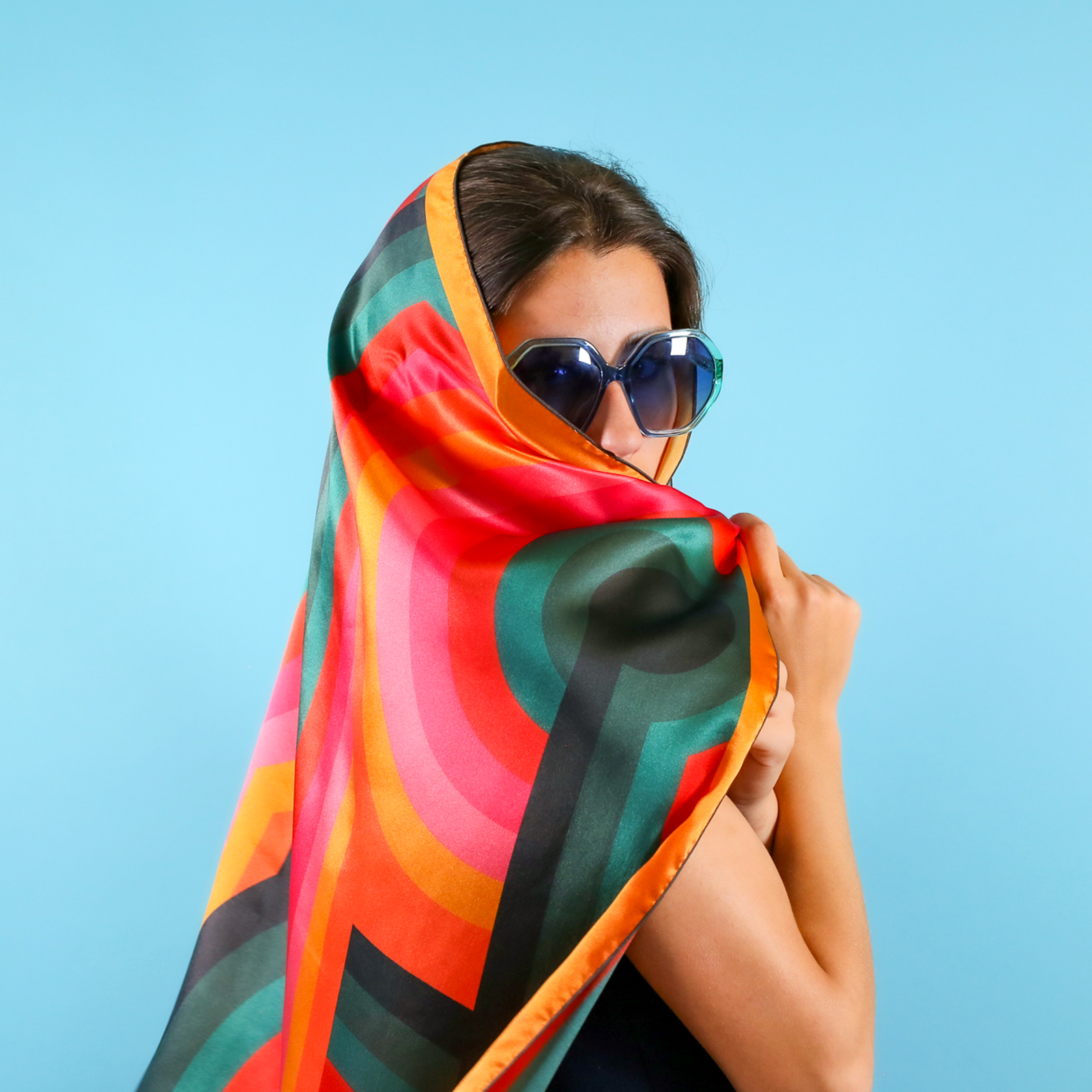 A woman wearing large blue sunglasses is partially covered with a vibrant, multicolored scarf against a light blue background.