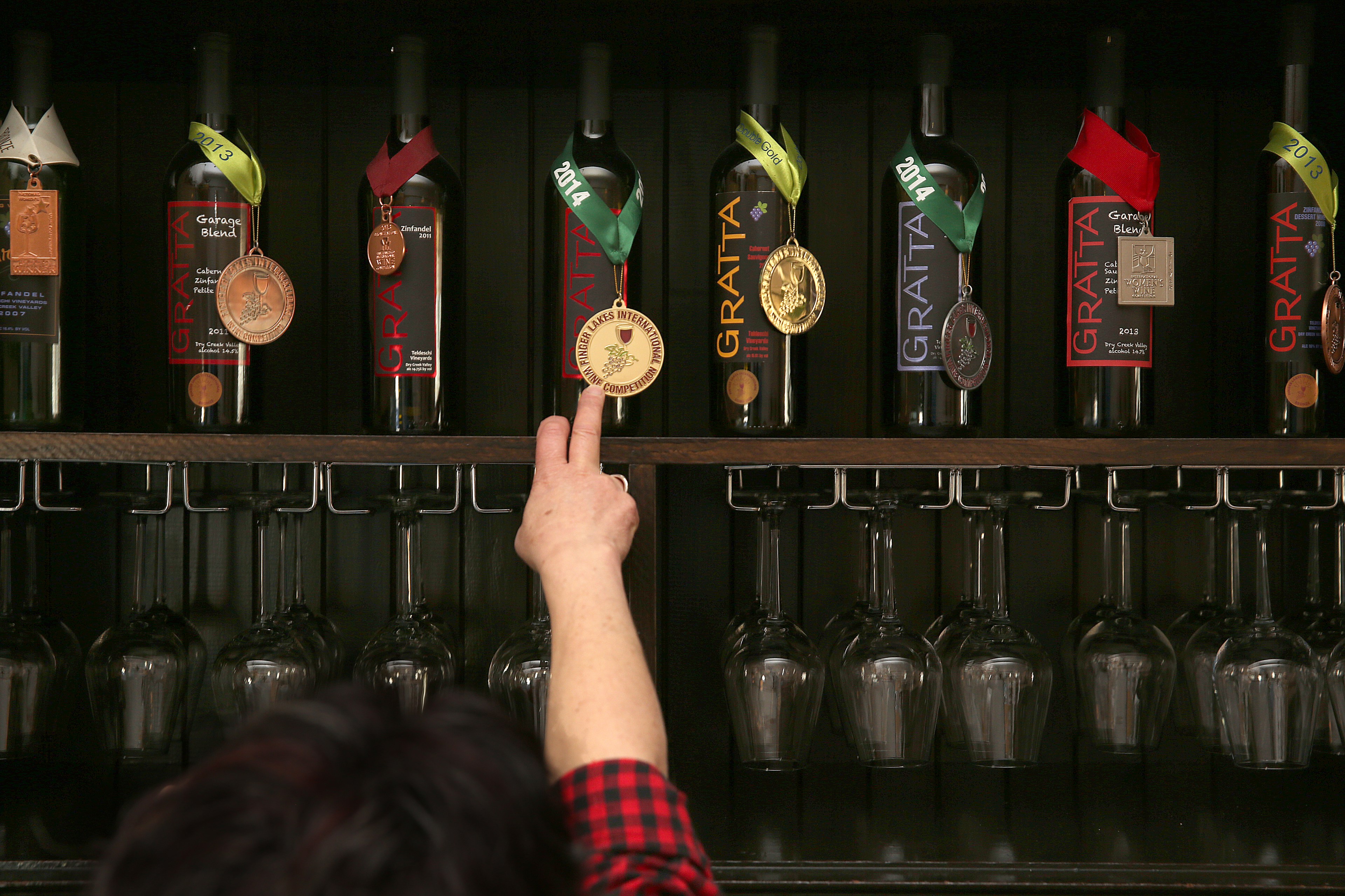 A hand points to a row of wine bottles, each adorned with medals. Below the bottles, wine glasses hang upside down on a rack.