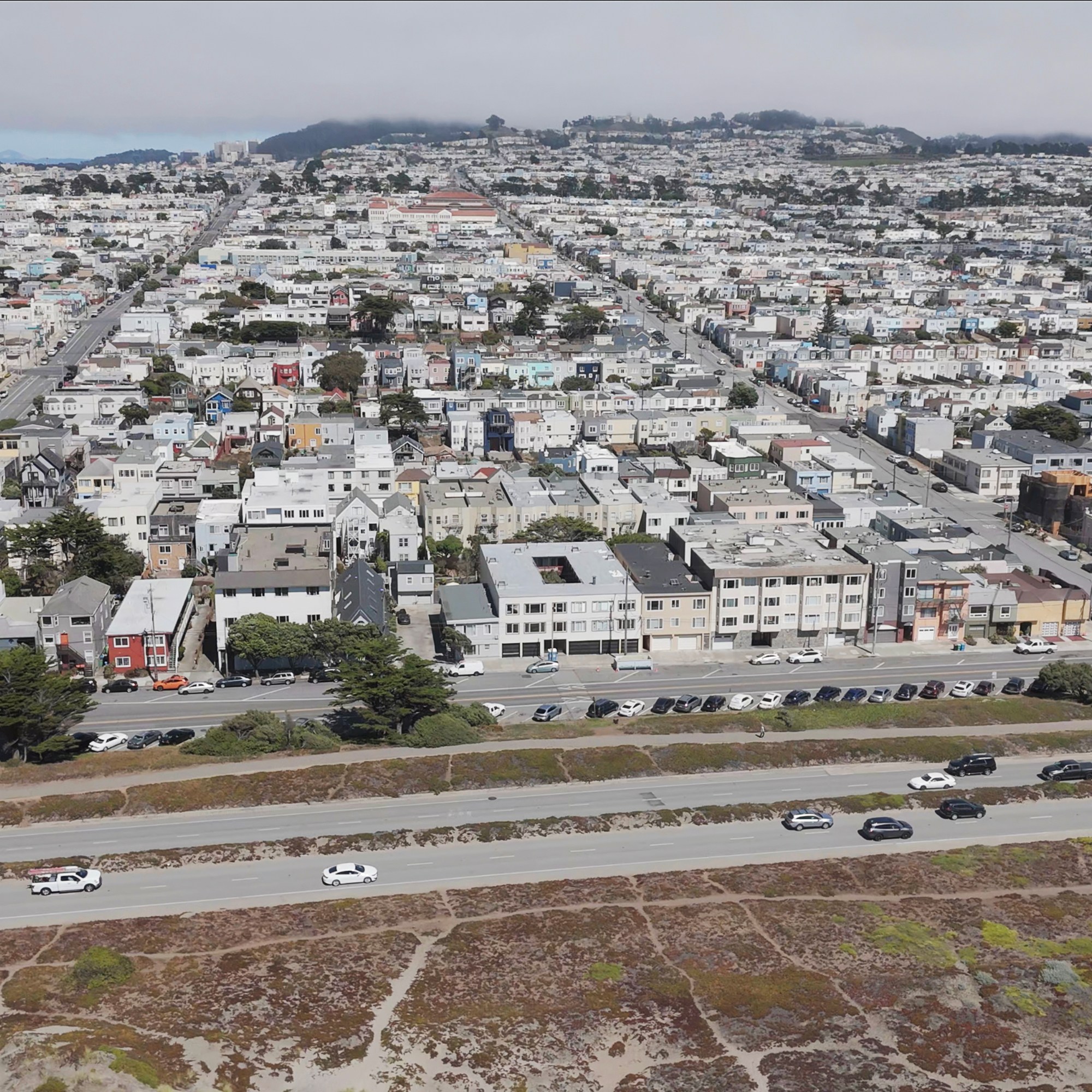 The image shows a densely packed suburban area with rows of houses, intersecting streets, and a highway with cars in the foreground. A hilly landscape is in the background.