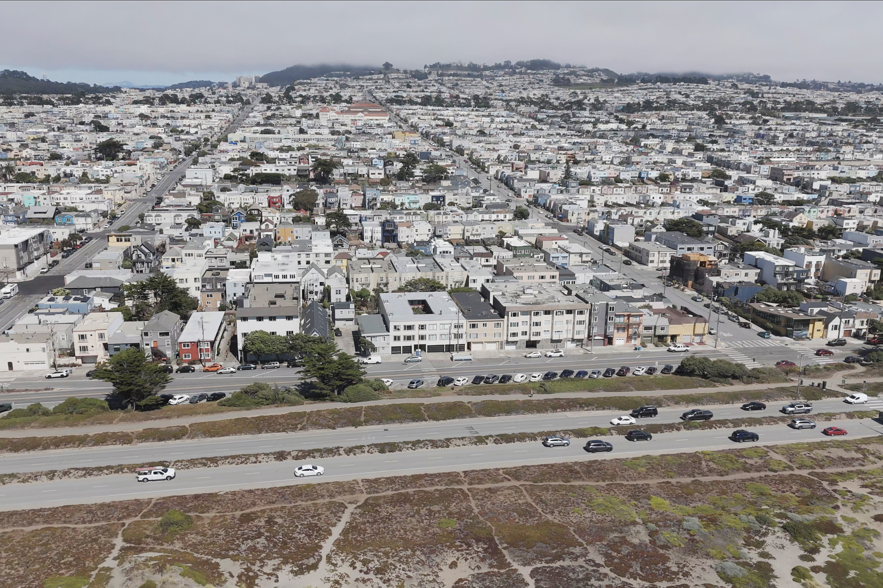 The image shows a densely packed suburban area with rows of houses, intersecting streets, and a highway with cars in the foreground. A hilly landscape is in the background.