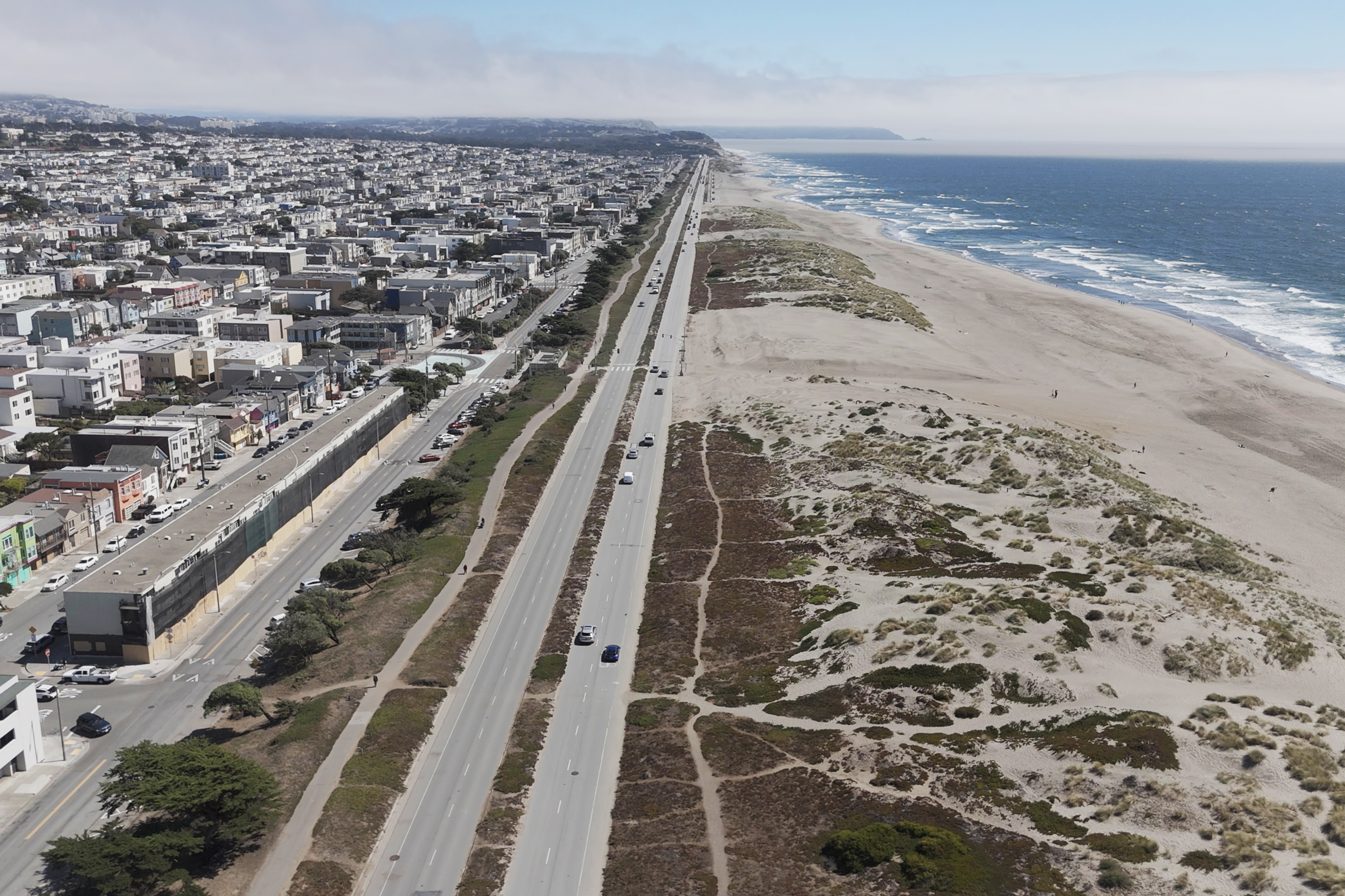 The image shows a coastal city with a wide beach on the right, a long road in the middle, and tightly packed residential buildings on the left. Waves crash on the shore.