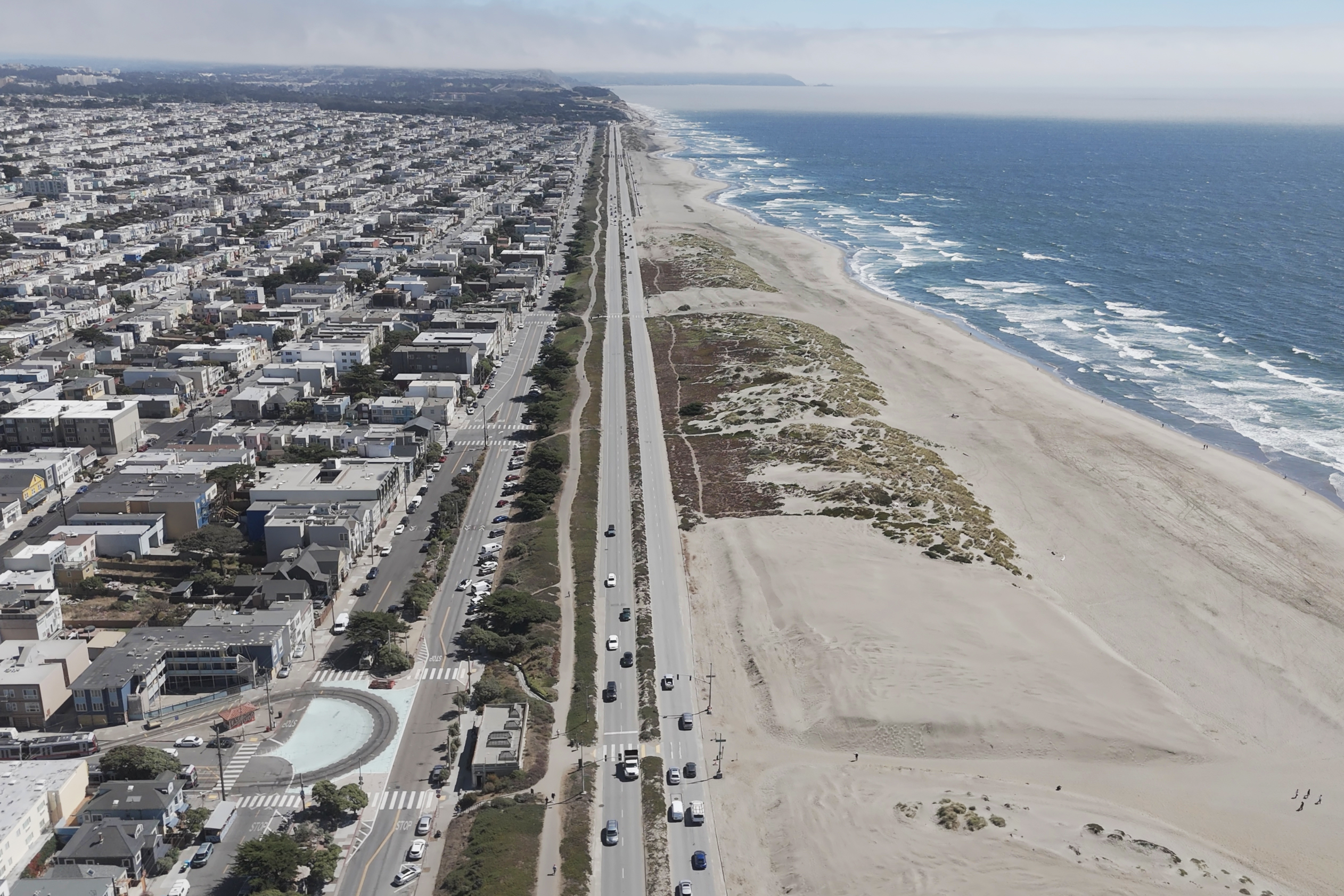 A coastal cityscape shows a dense urban area on the left, bordered by a highway, with a wide sandy beach and blue ocean waves on the right.