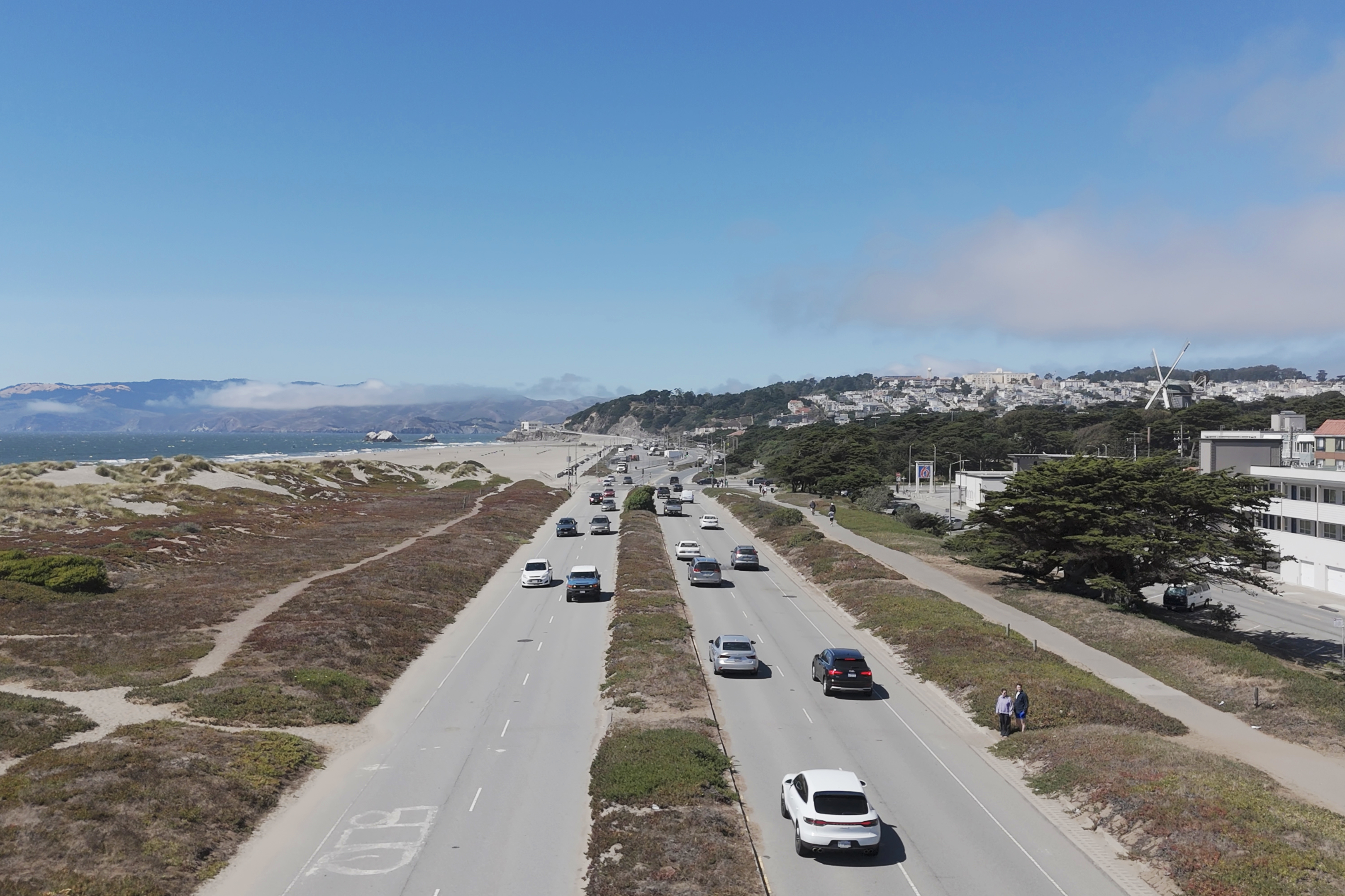 The image shows a road with cars driving between dunes and greenery. A clear blue sky and distant hills meet a cityscape, featuring modern buildings and a large windmill.