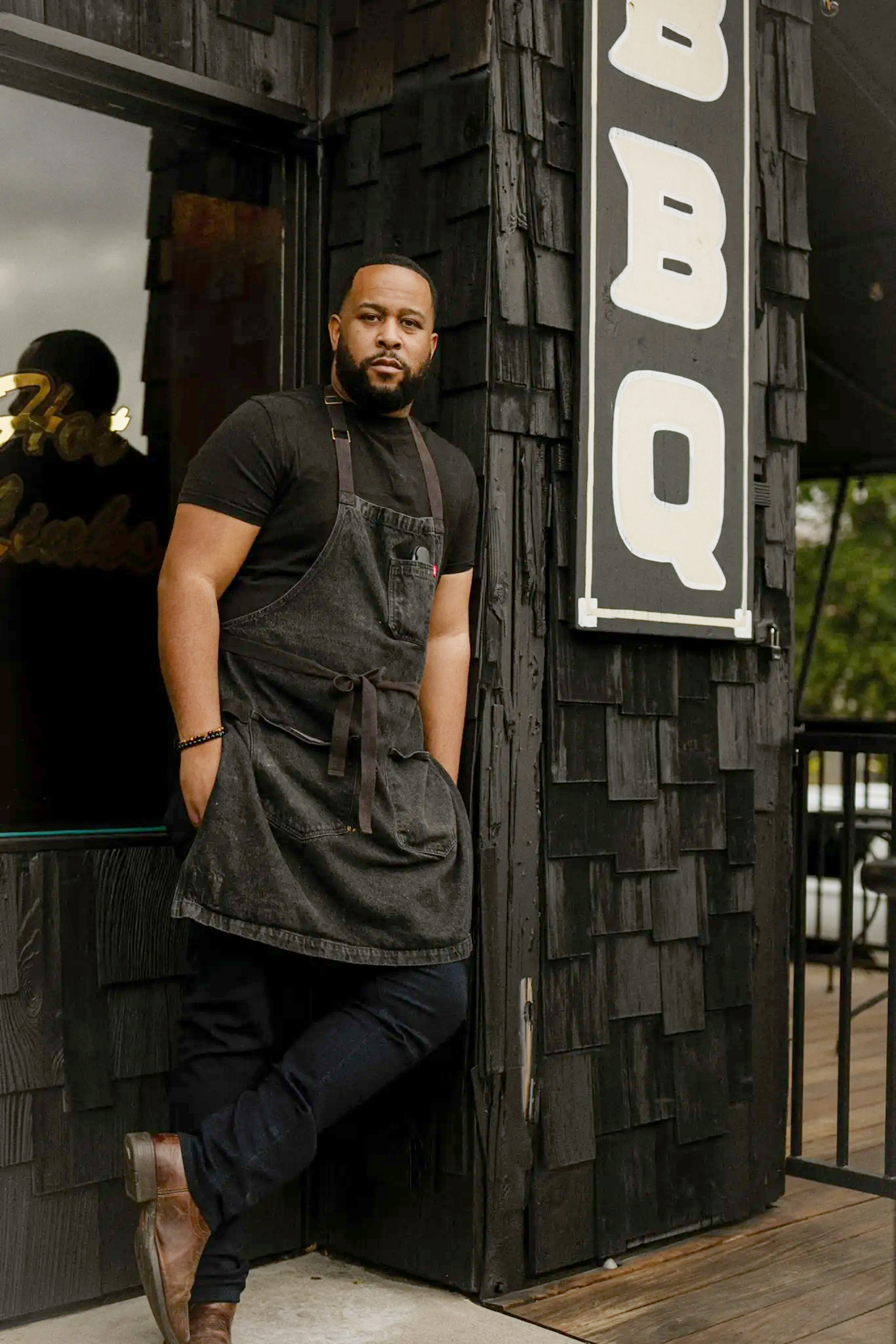 A man in a black apron stands confidently outside a BBQ restaurant. He's leaning against a building with black wood siding and a large "BBQ" sign.