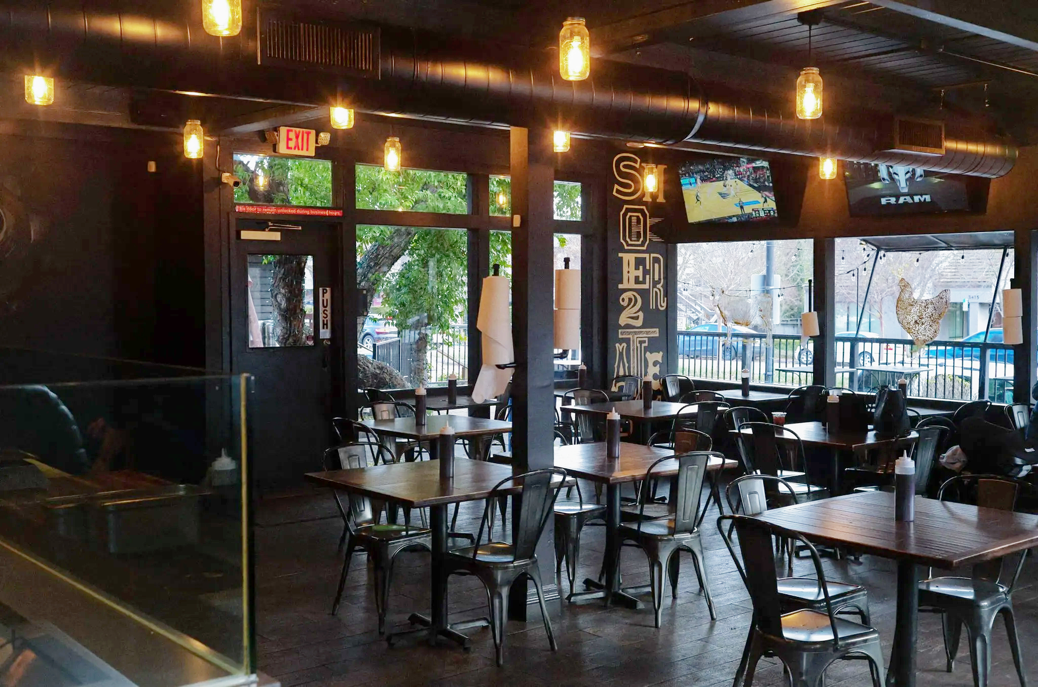 The image shows a dimly lit restaurant with metal chairs, wooden tables, and hanging lights. There are two TVs, a wall with decorative text, and a window view outside.