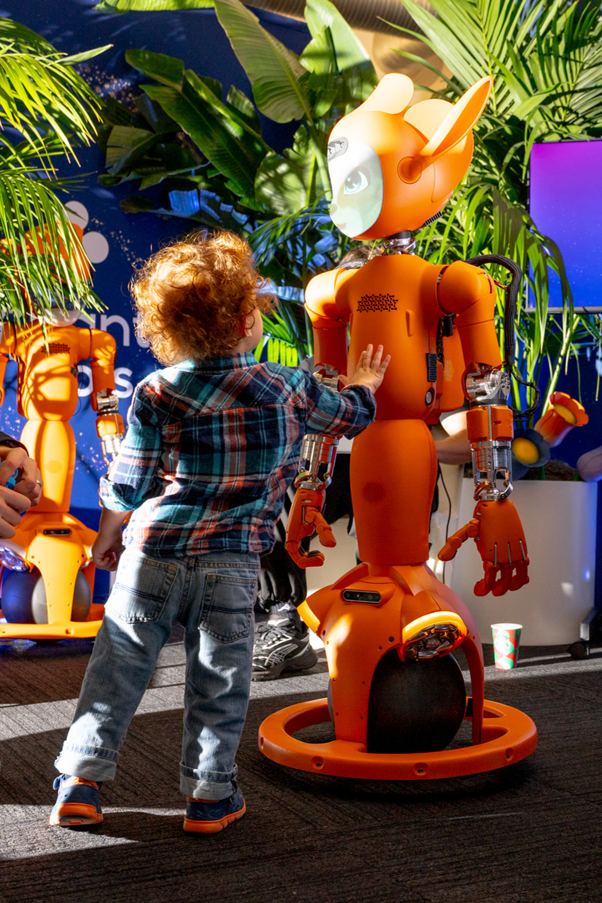 A young child in a plaid shirt touches a bright orange robot surrounded by lush green plants, creating a playful and futuristic scene.