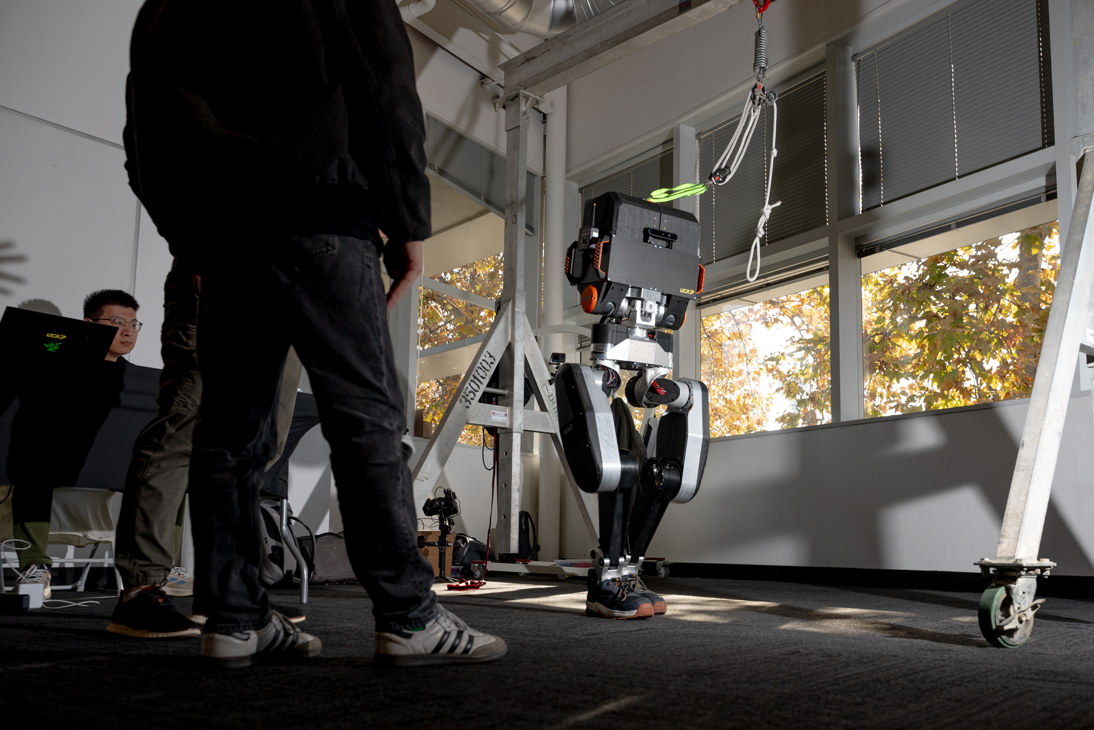 A humanoid robot with black and silver legs stands under a support structure indoors. Two people nearby observe it, one with a laptop, amid autumnal light.