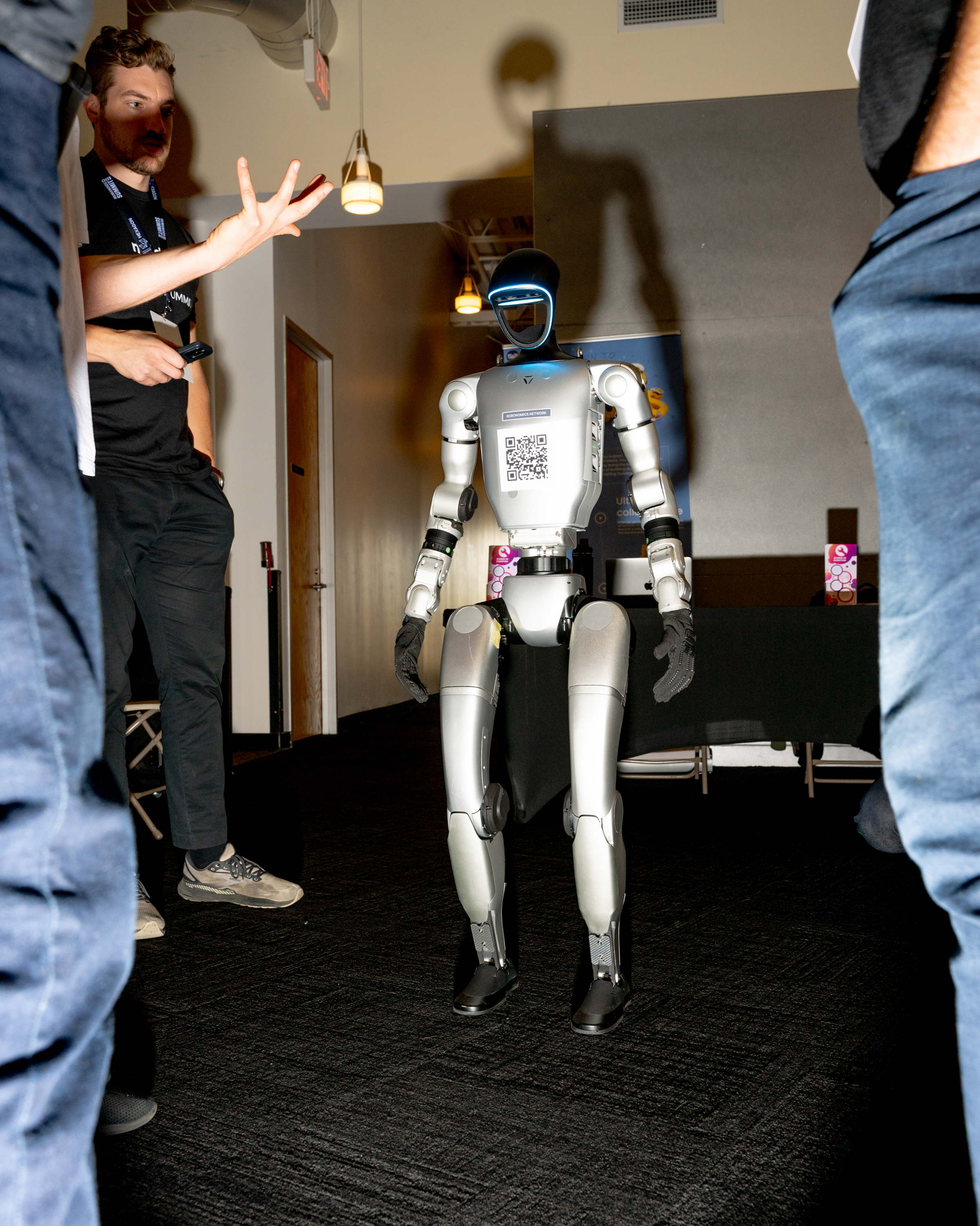 A humanoid robot stands in a room with people around it. The robot has a QR code on its chest plate and appears to be the focus of attention.