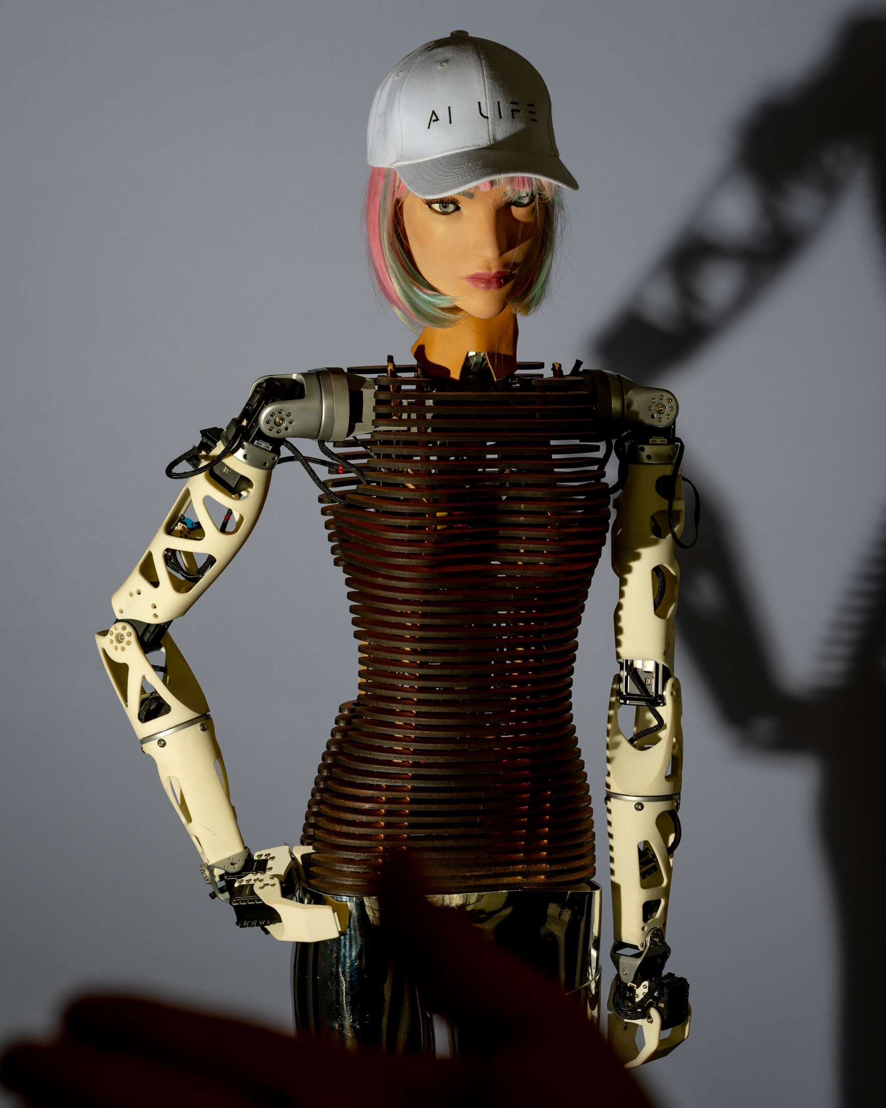 A humanoid robot with a textured torsoless body and mechanical arms wears a white &quot;AI LIFE&quot; cap. It has colorful hair and casts a shadow on the wall.