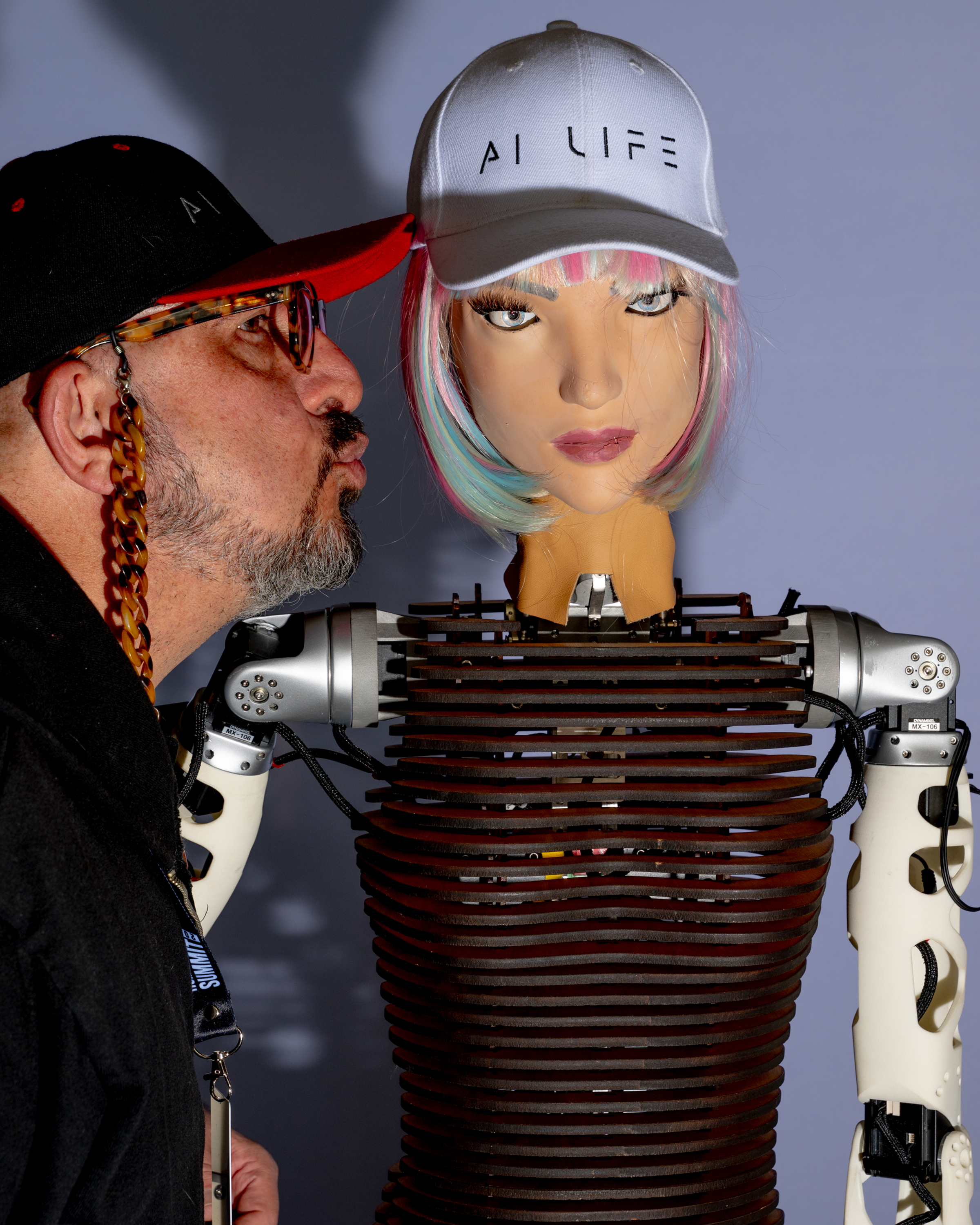 A man wearing glasses and a cap leans in to kiss a humanoid robot with colorful hair, both wearing caps labeled "AI LIFE." The robot has a visible mechanical body.