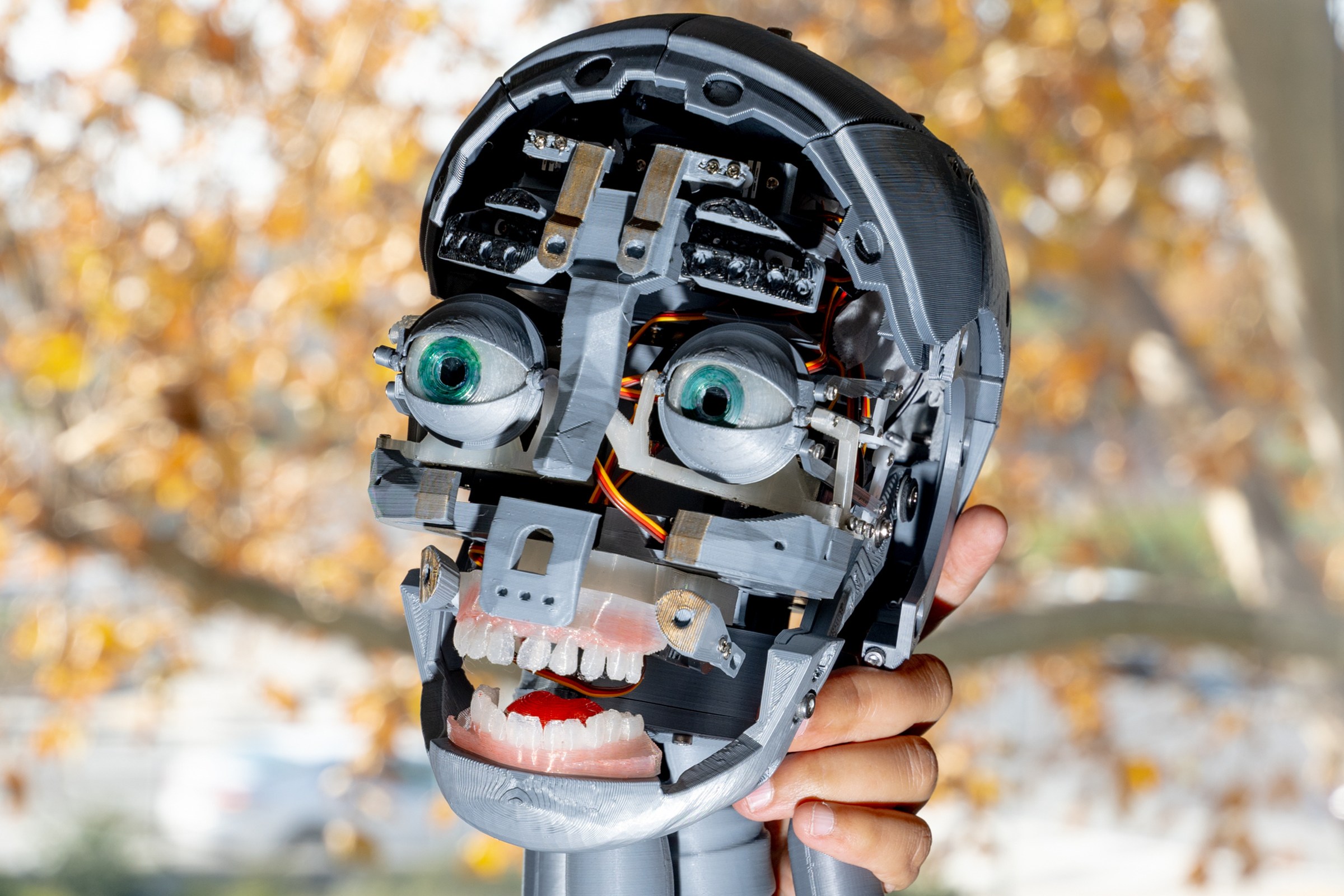 A person holds a robotic head with exposed mechanical components, large green eyes, and artificial teeth, set against a background of autumn trees.