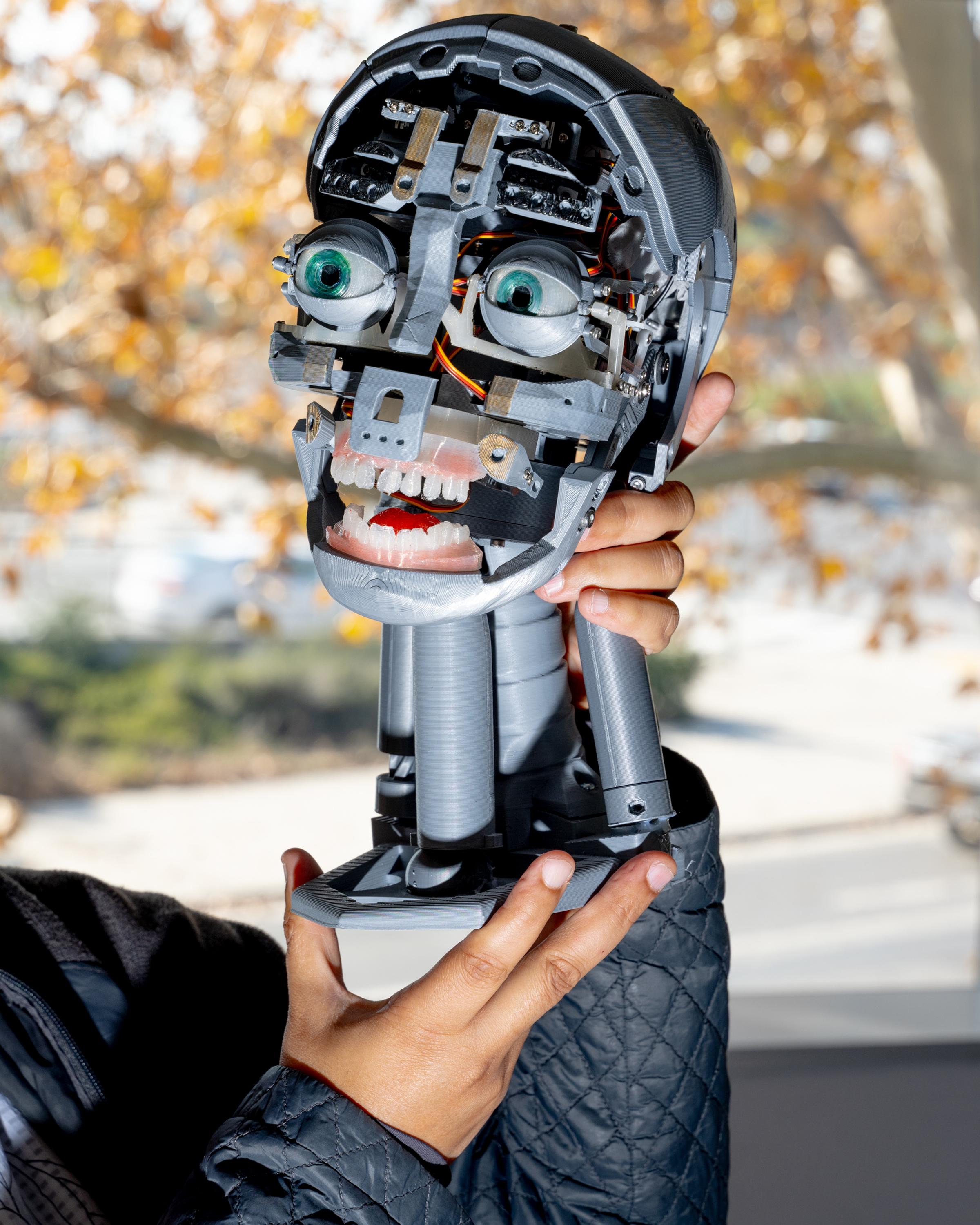 A person holds a robotic head with exposed mechanical components, large green eyes, and artificial teeth, set against a background of autumn trees.