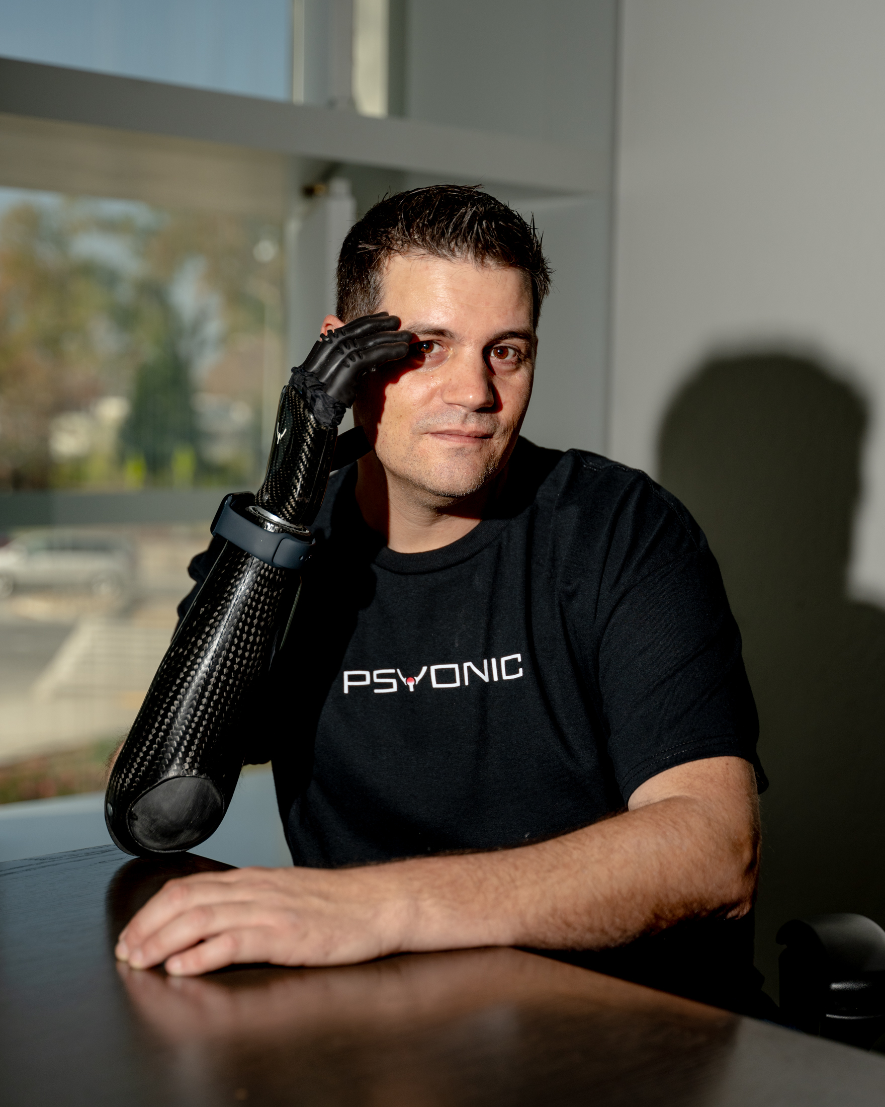 A man wearing a black shirt with the word &quot;PSYONIC&quot; is sitting at a table. He is resting his chin on his hand, which is part of a sleek, black prosthetic arm.