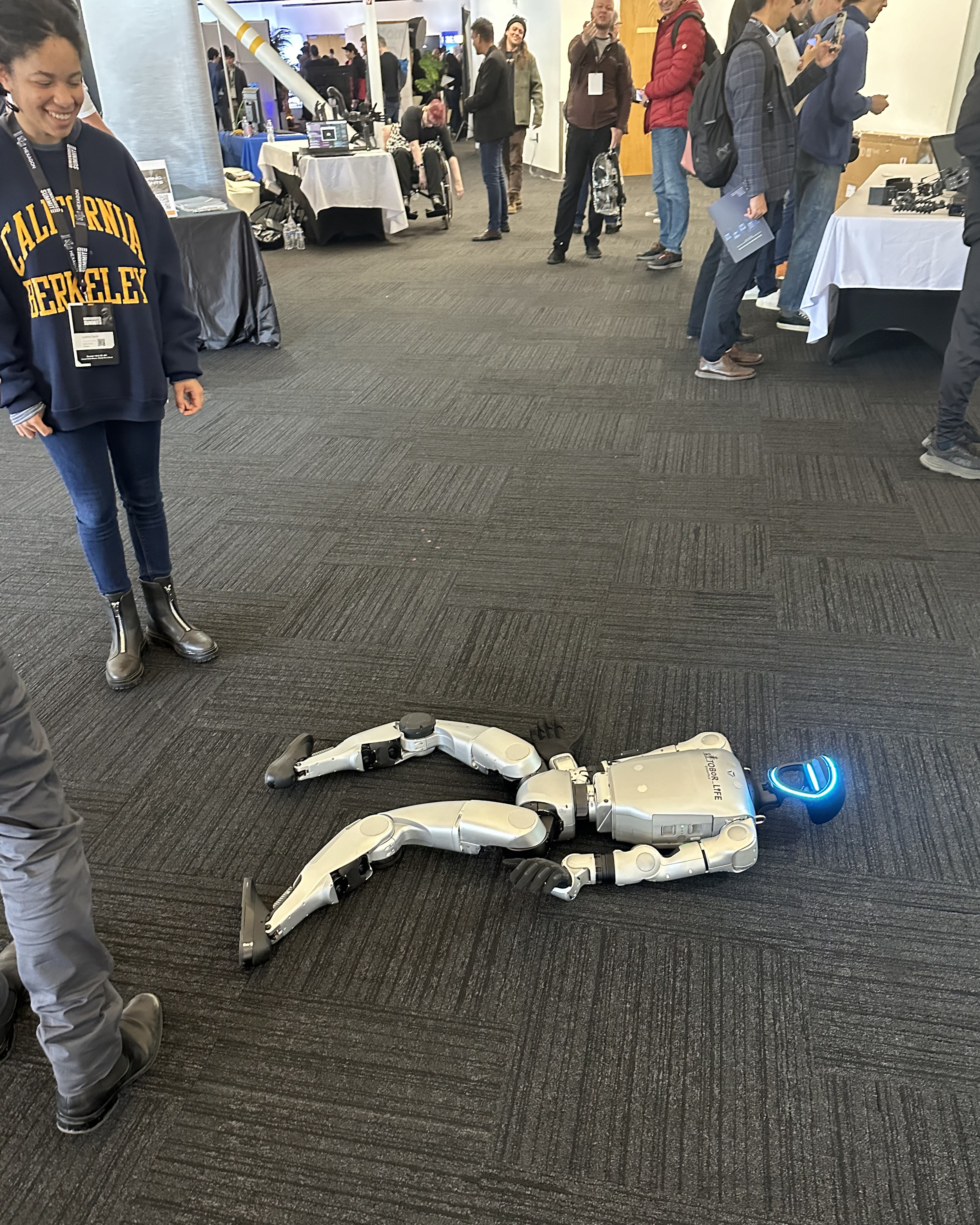 A humanoid robot lies on the floor with a glowing blue face, surrounded by people casually observing. One person is wearing a &quot;California Berkeley&quot; sweatshirt.