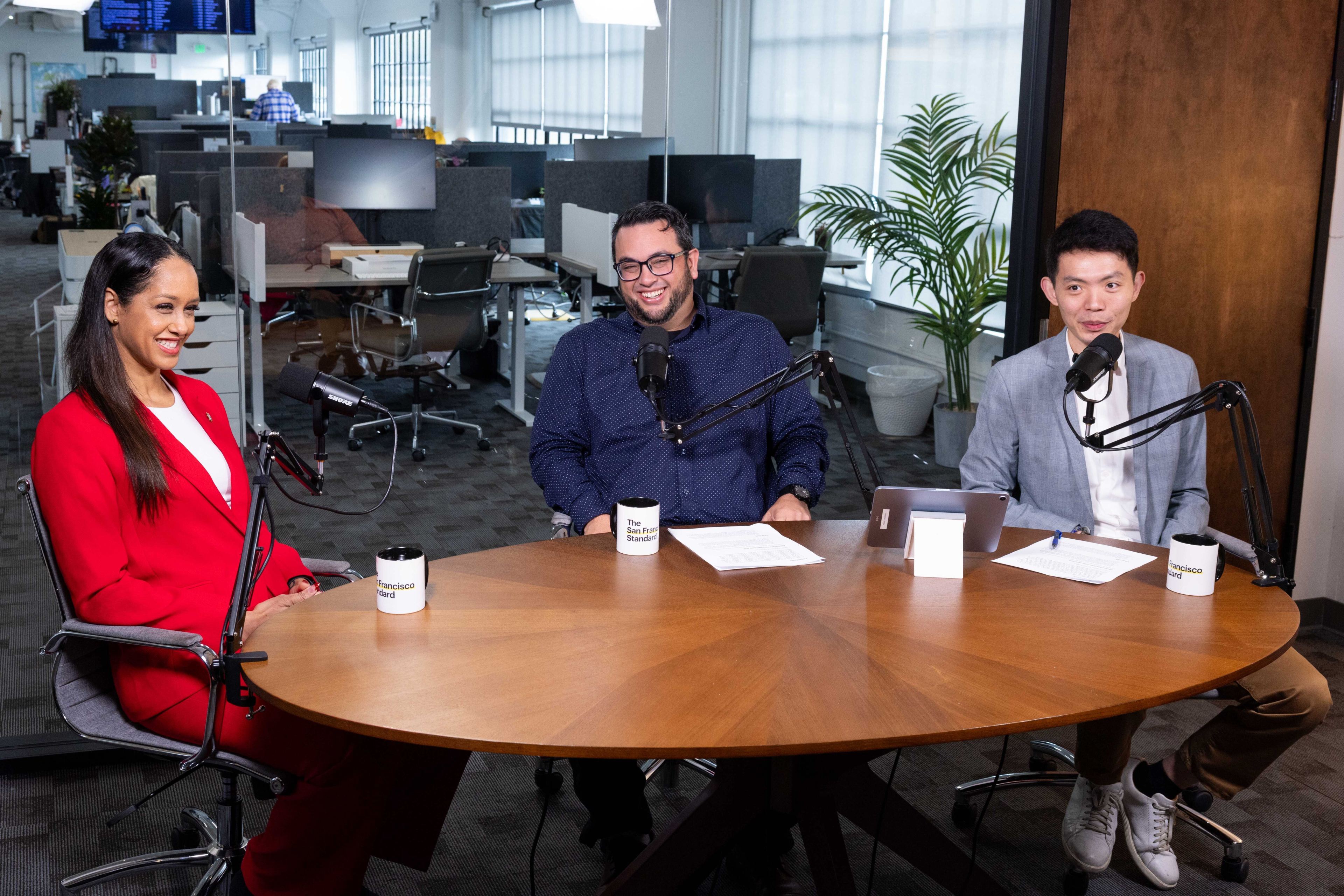 Brooke Jenkins in a red suit seated, left, at a table with two men reporters in blue and gray. 