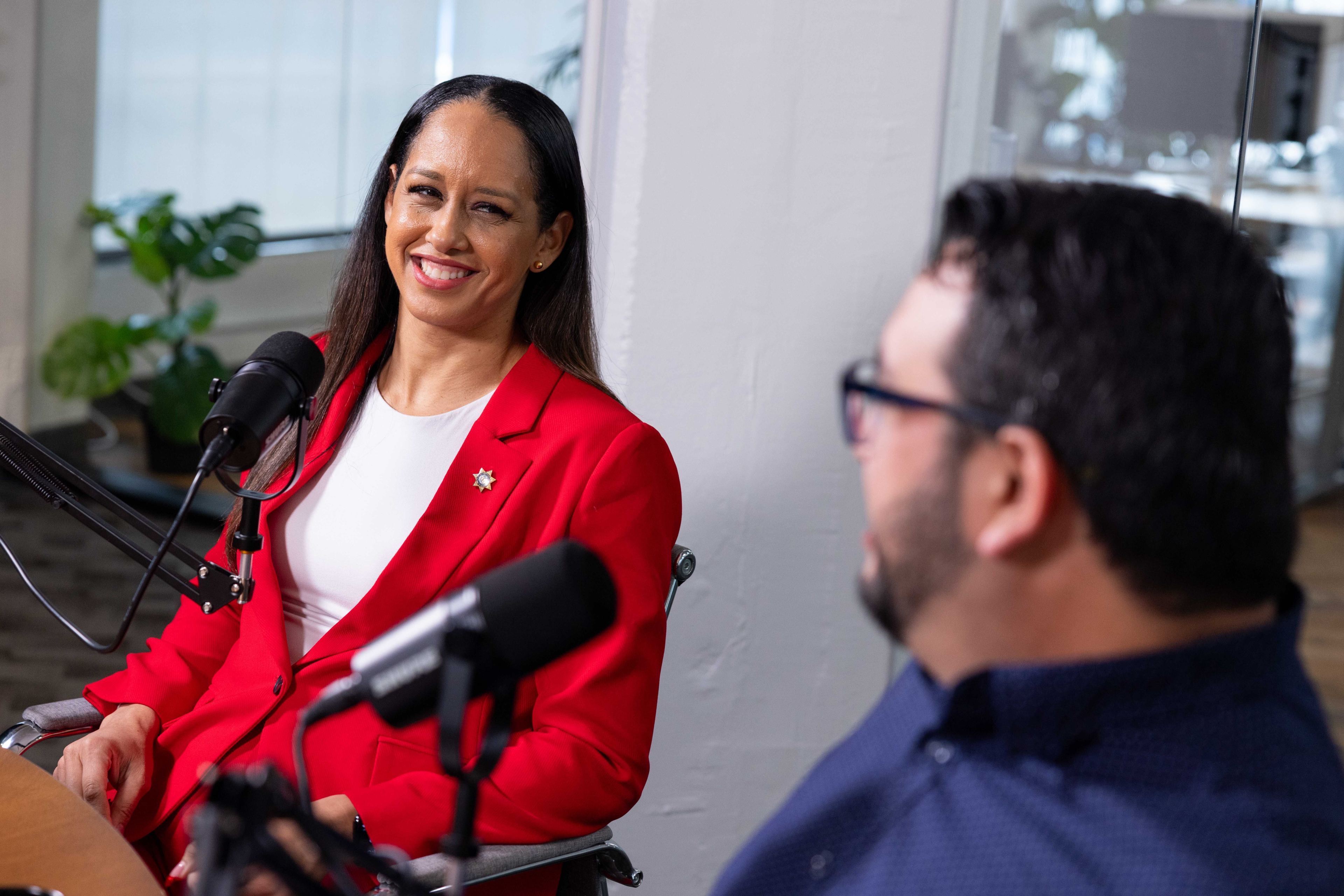 A woman in a red suit on the left, seated, smiling at a table with microphones.