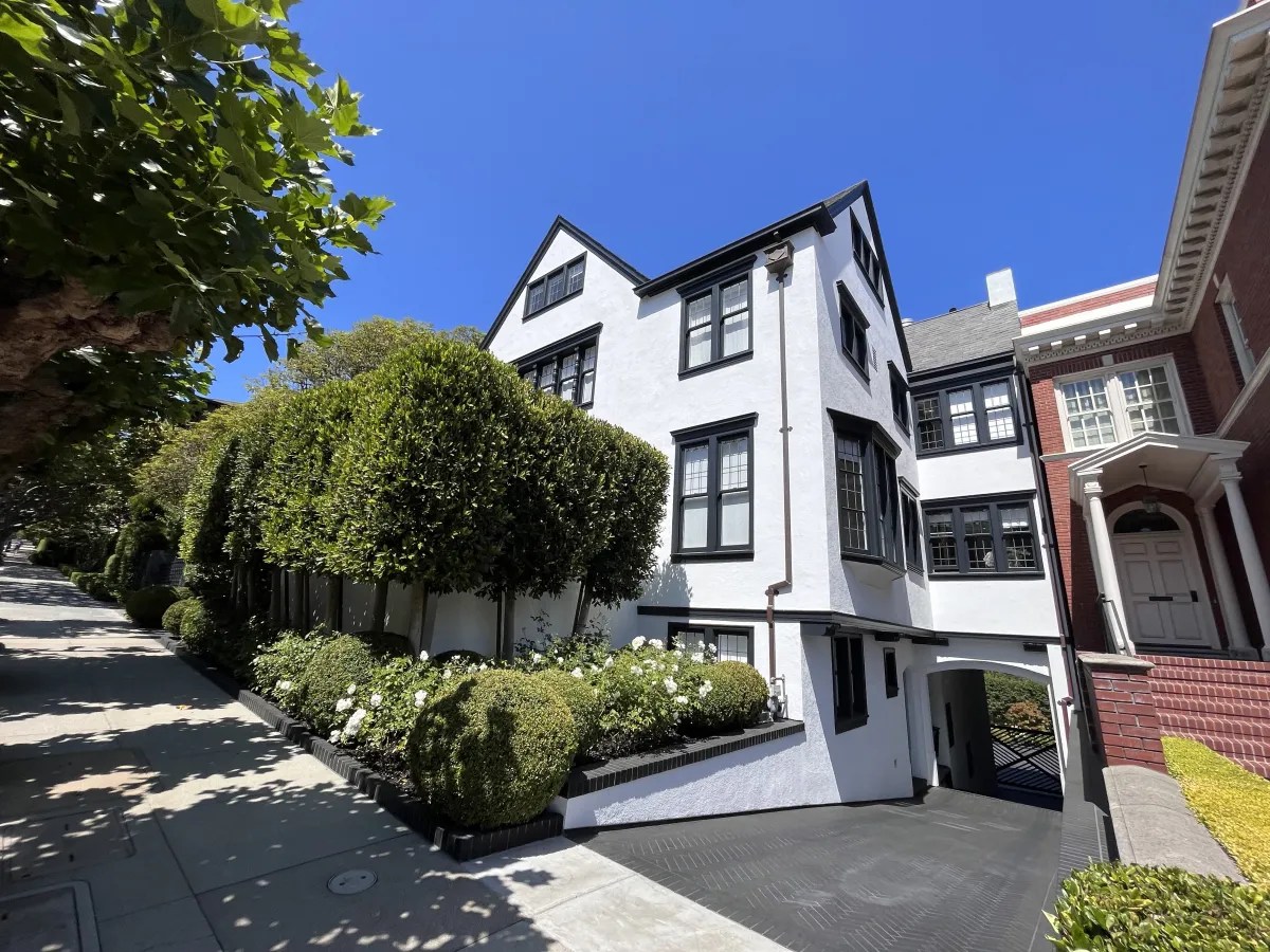 A white multi-story house with black trim sits on a hilly street. Manicured hedges and bushes line the sidewalk, and a blue sky is overhead.