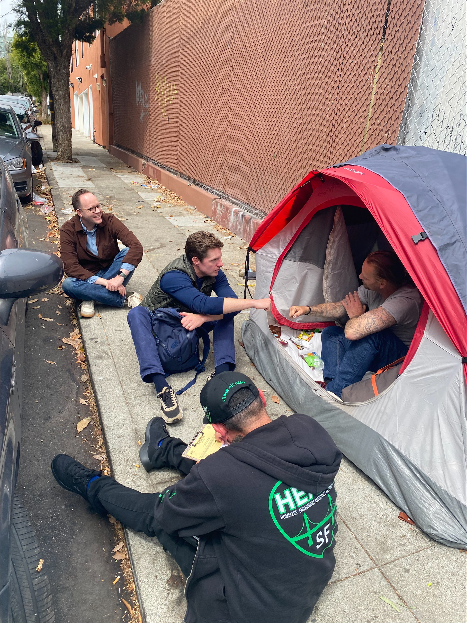 Three men sit on a sidewalk next to a tent, engaged in conversation. Two are outside the tent, one is inside. Fallen leaves line the street beside parked cars.