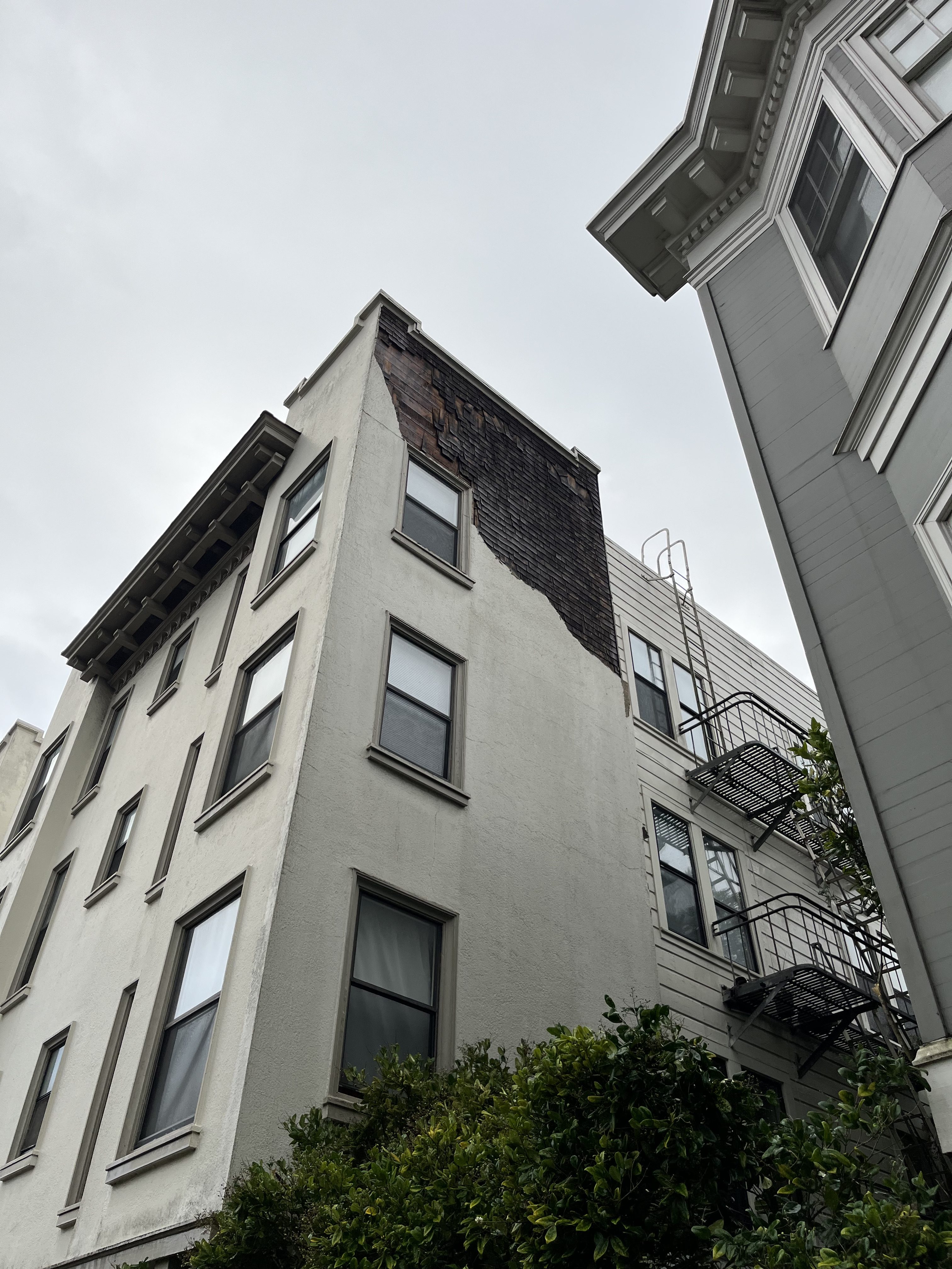 A four-story building with an exposed wooden section on its corner contrasts with its predominantly white facade. Adjacent fire escapes and a tree are visible.