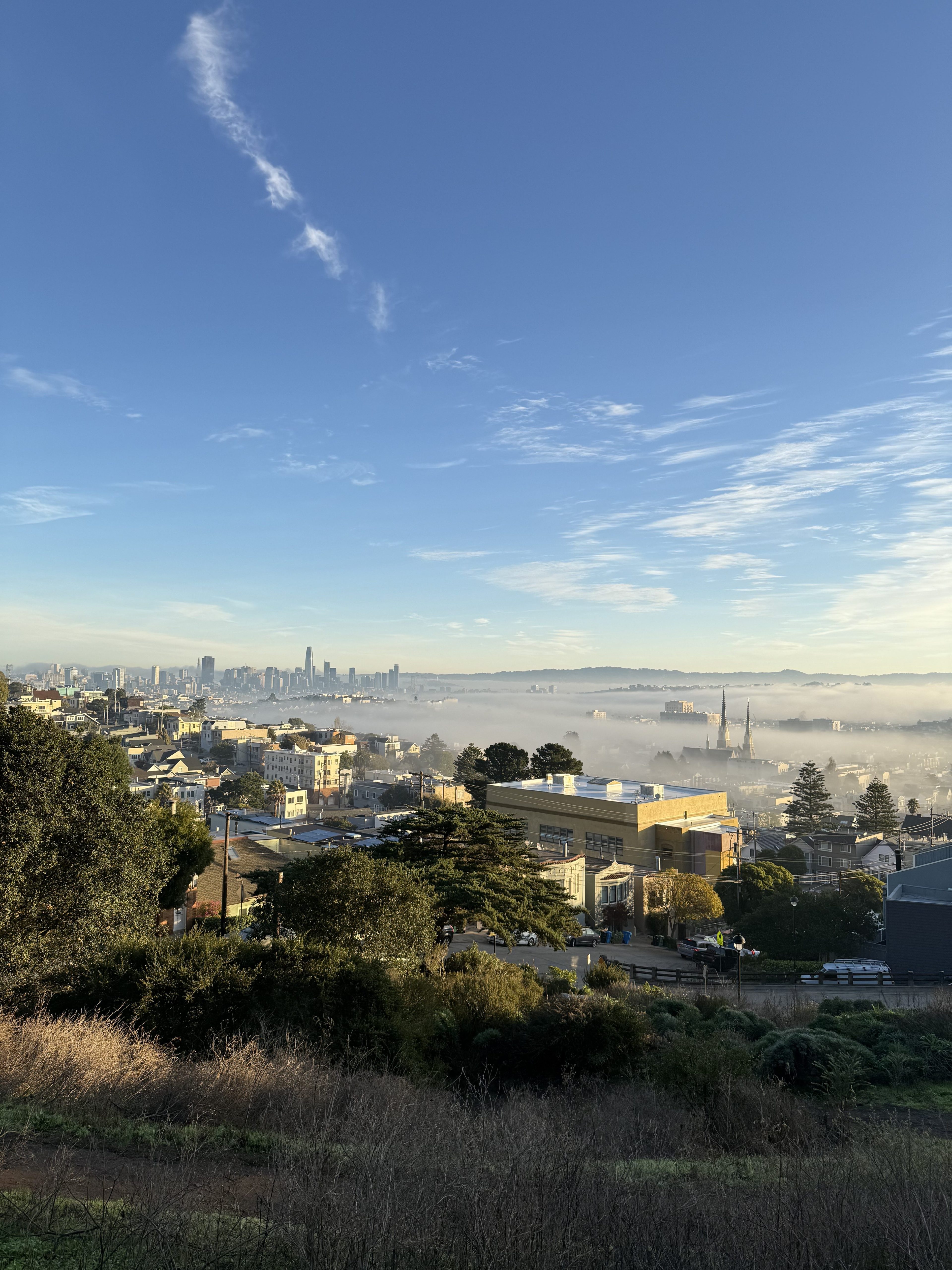 A cityscape view shows a sprawling city partially covered in fog. Tall buildings are visible in the distance, and the sky is clear with scattered clouds above.