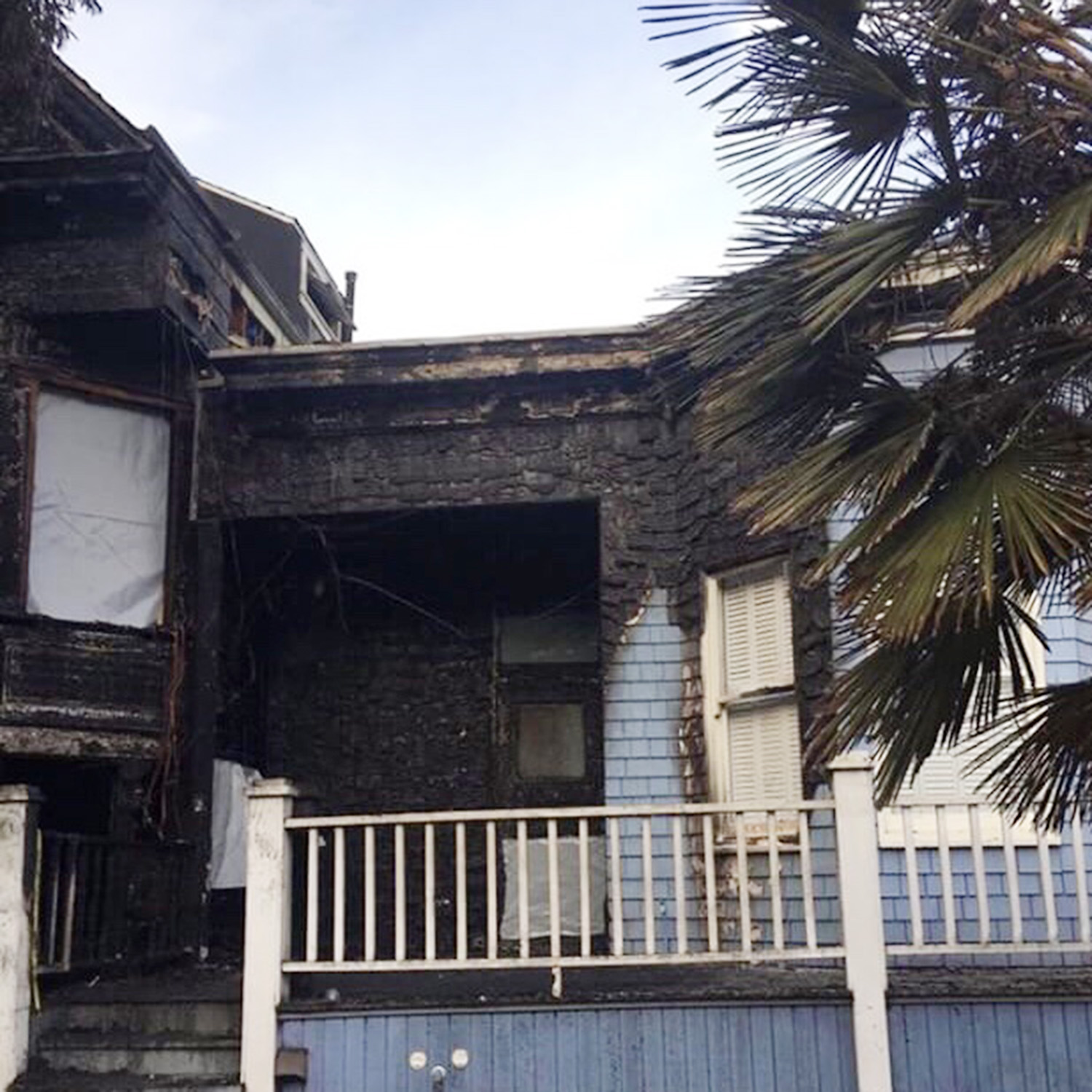 The image shows a charred and burned two-story house with visible fire damage. There's a palm tree on the right side, and the sky is clear.