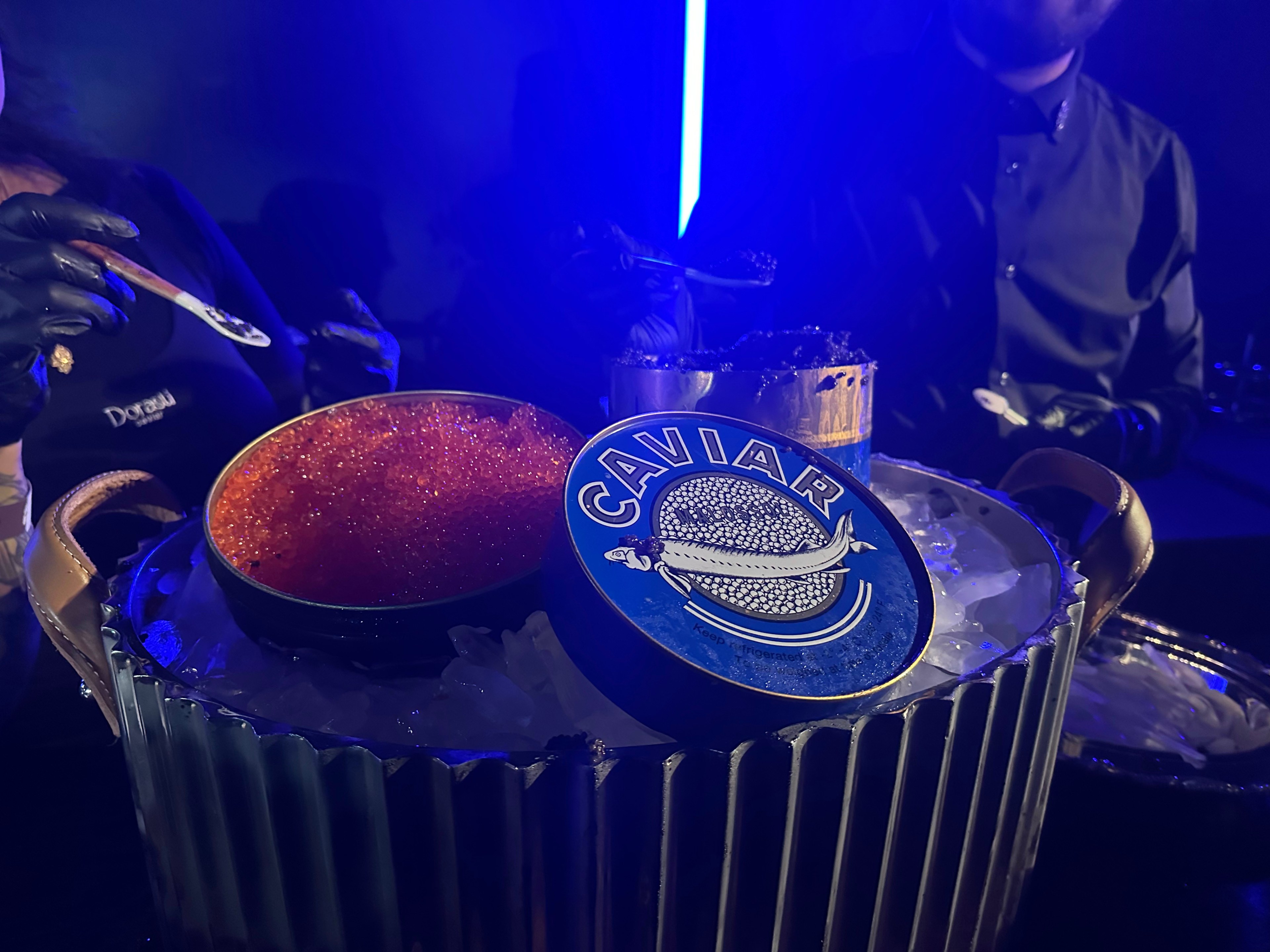 Two people serve caviar from an elegant container filled with ice. There's a spotlight highlighting the colorful caviar and a decorative lid nearby.