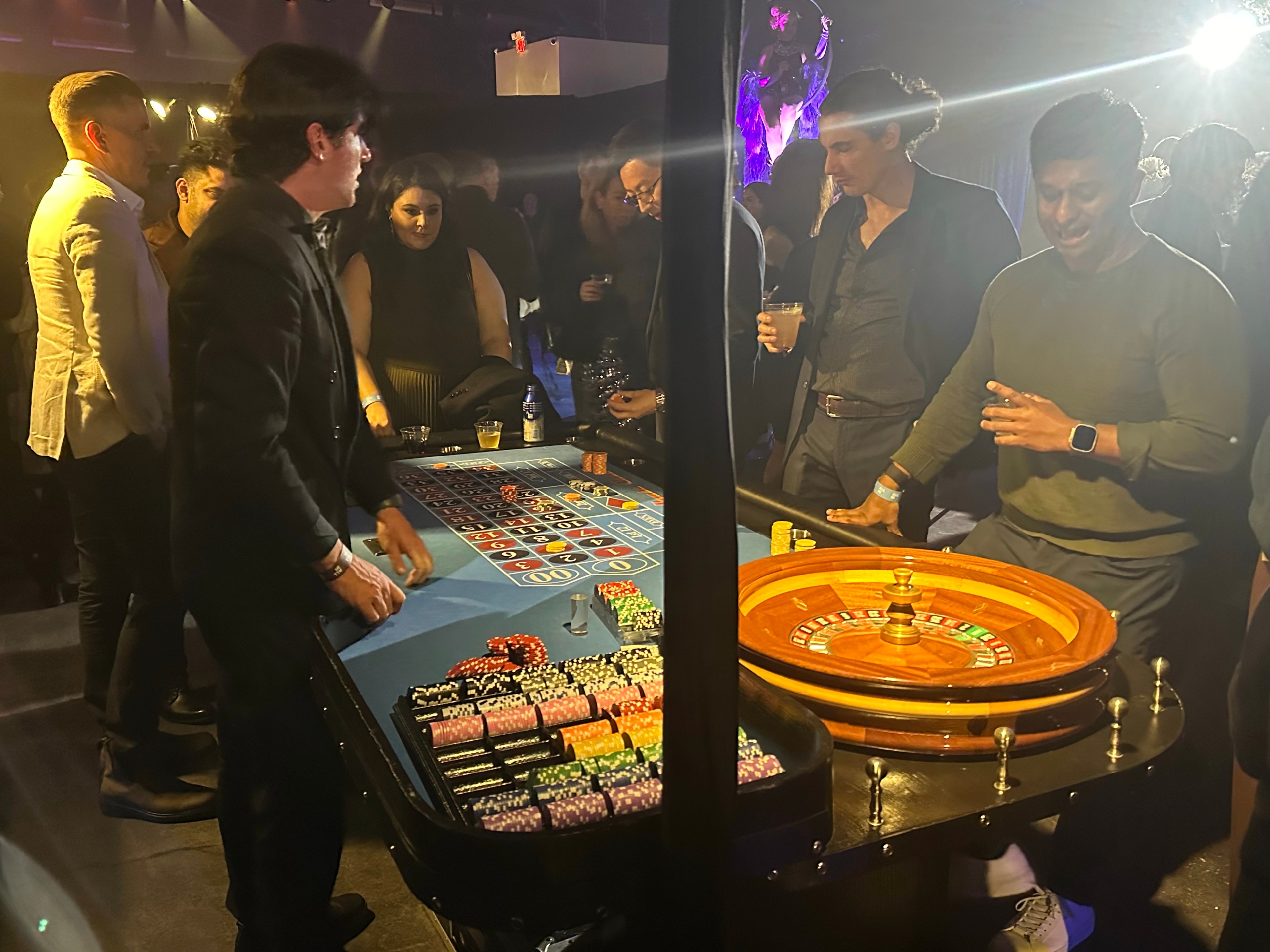 People are gathered around a roulette table in a dimly lit room. The roulette wheel is in action, and guests, dressed in formal attire, are engaged in the game.