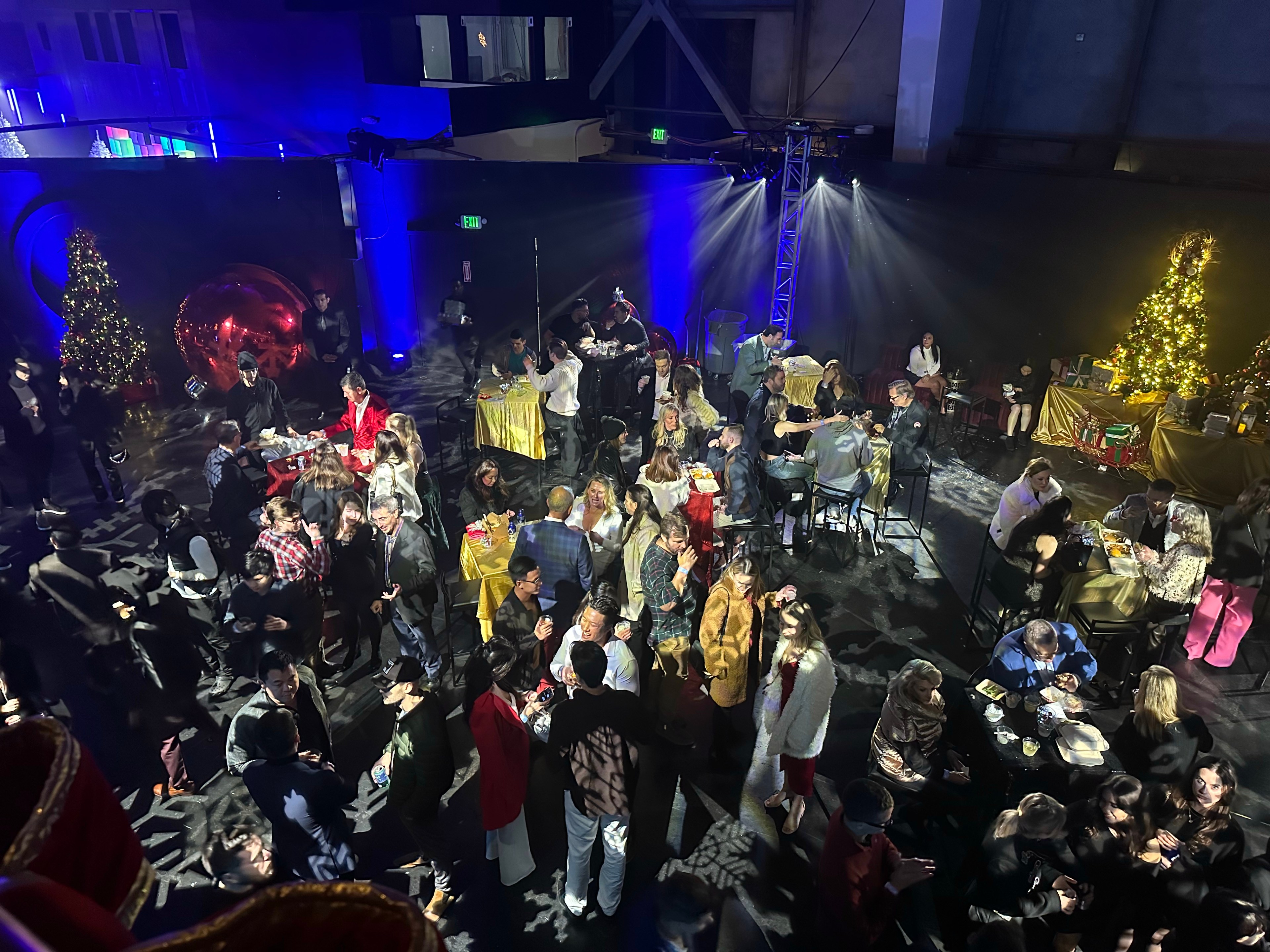 The image shows a festive party with people mingling around tables, decorated with Christmas trees and gifts. The lighting is colorful, creating a lively atmosphere.