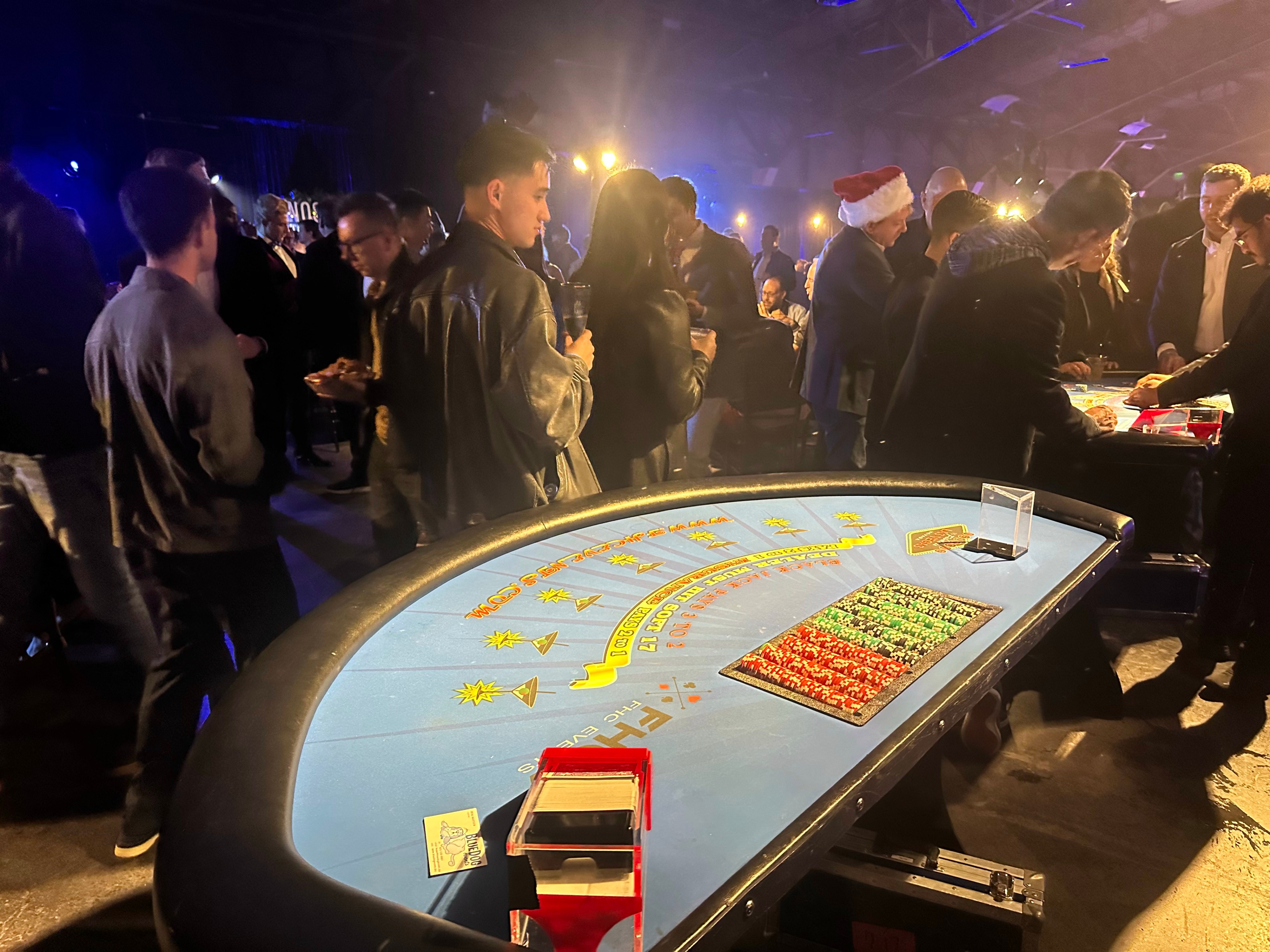 People are gathered around casino tables in a dimly lit room. Some are wearing festive attire, and there are chips and cards on the tables.