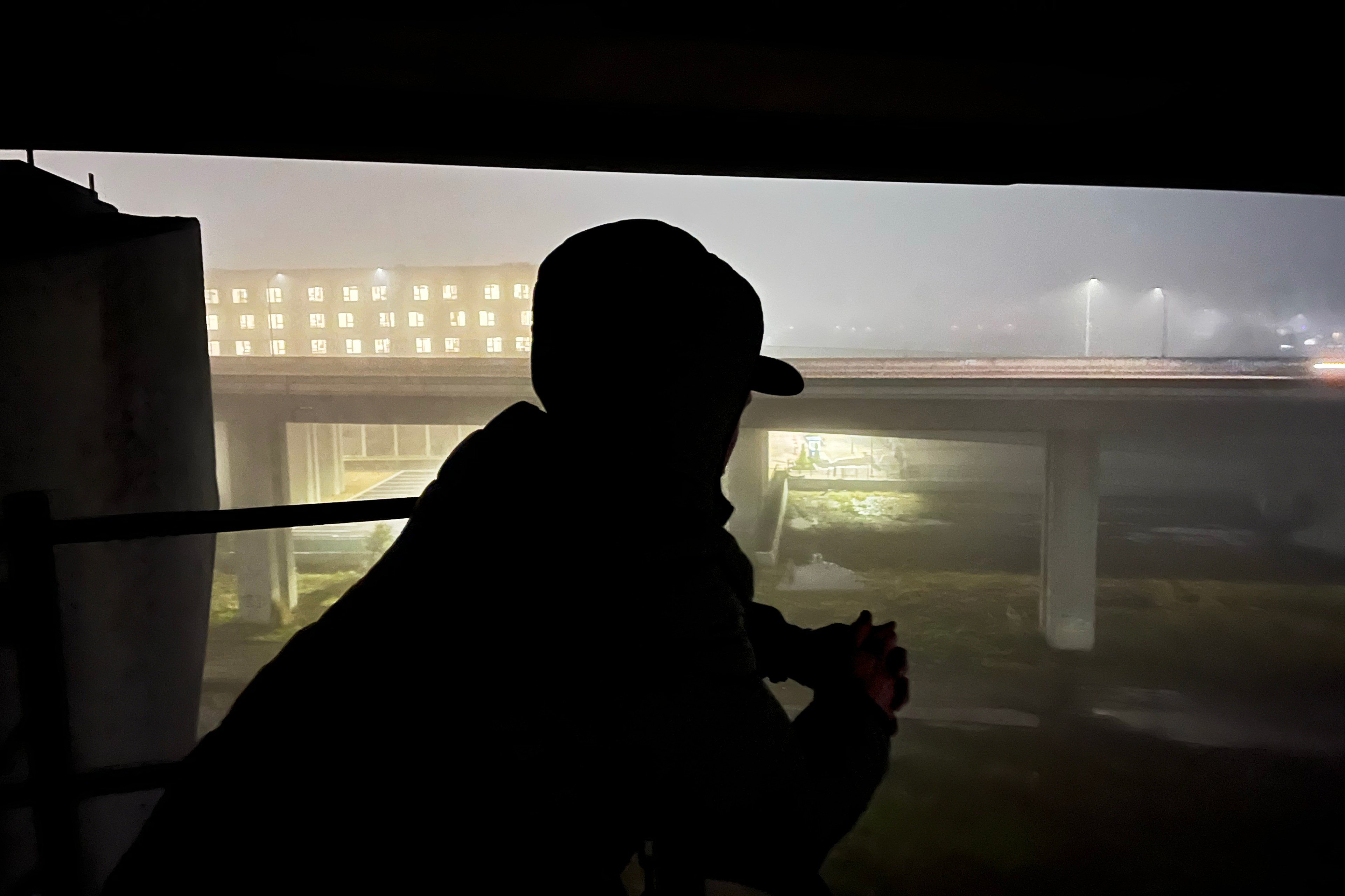 A silhouette of a person in a hood gazes out at a foggy urban scene, featuring a building with lit windows and an overpass with streetlights.