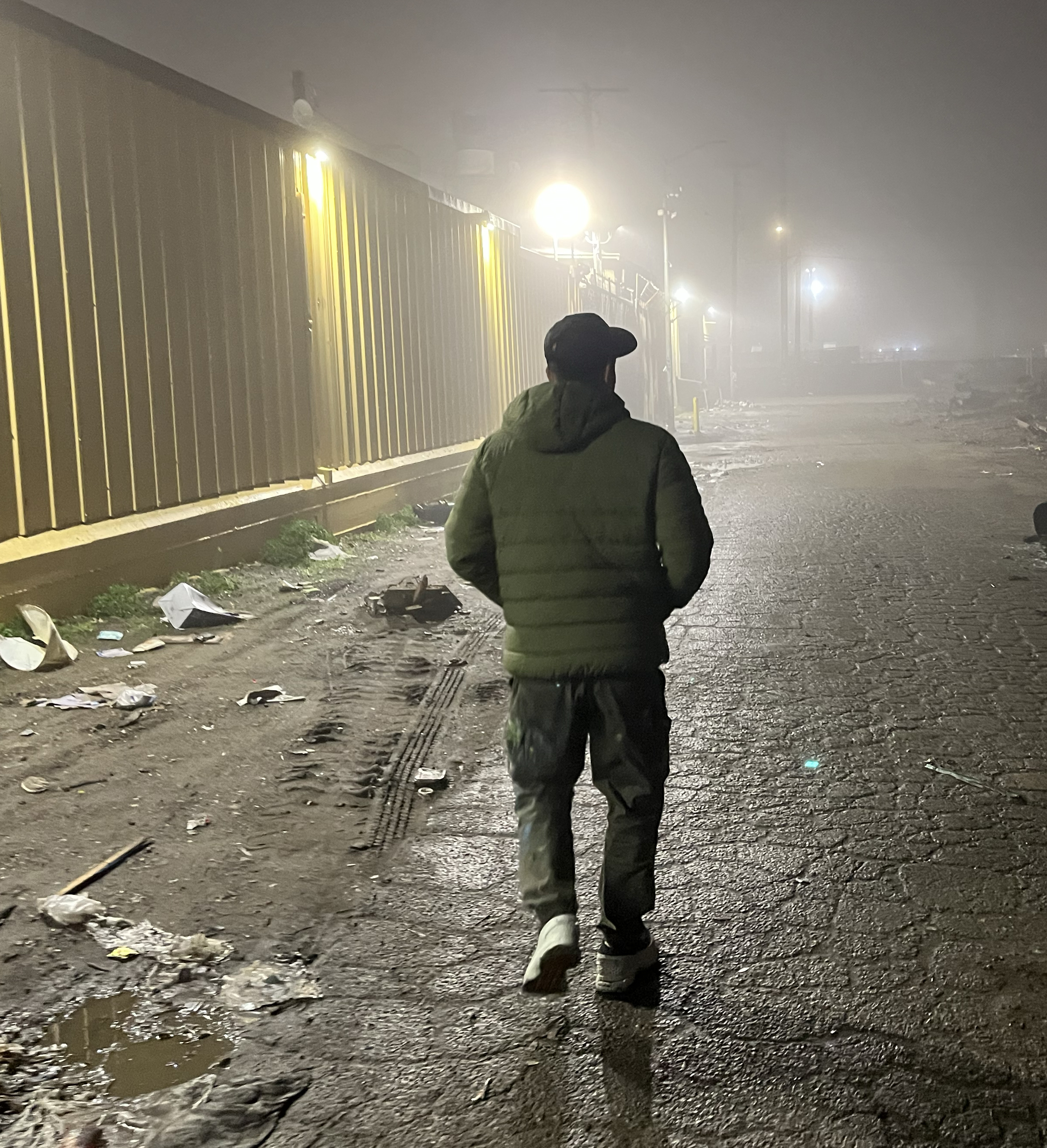 A person in a hooded jacket and cap walks on a dimly lit, foggy street. Trash is scattered on the ground near a long building with bright overhead lights.