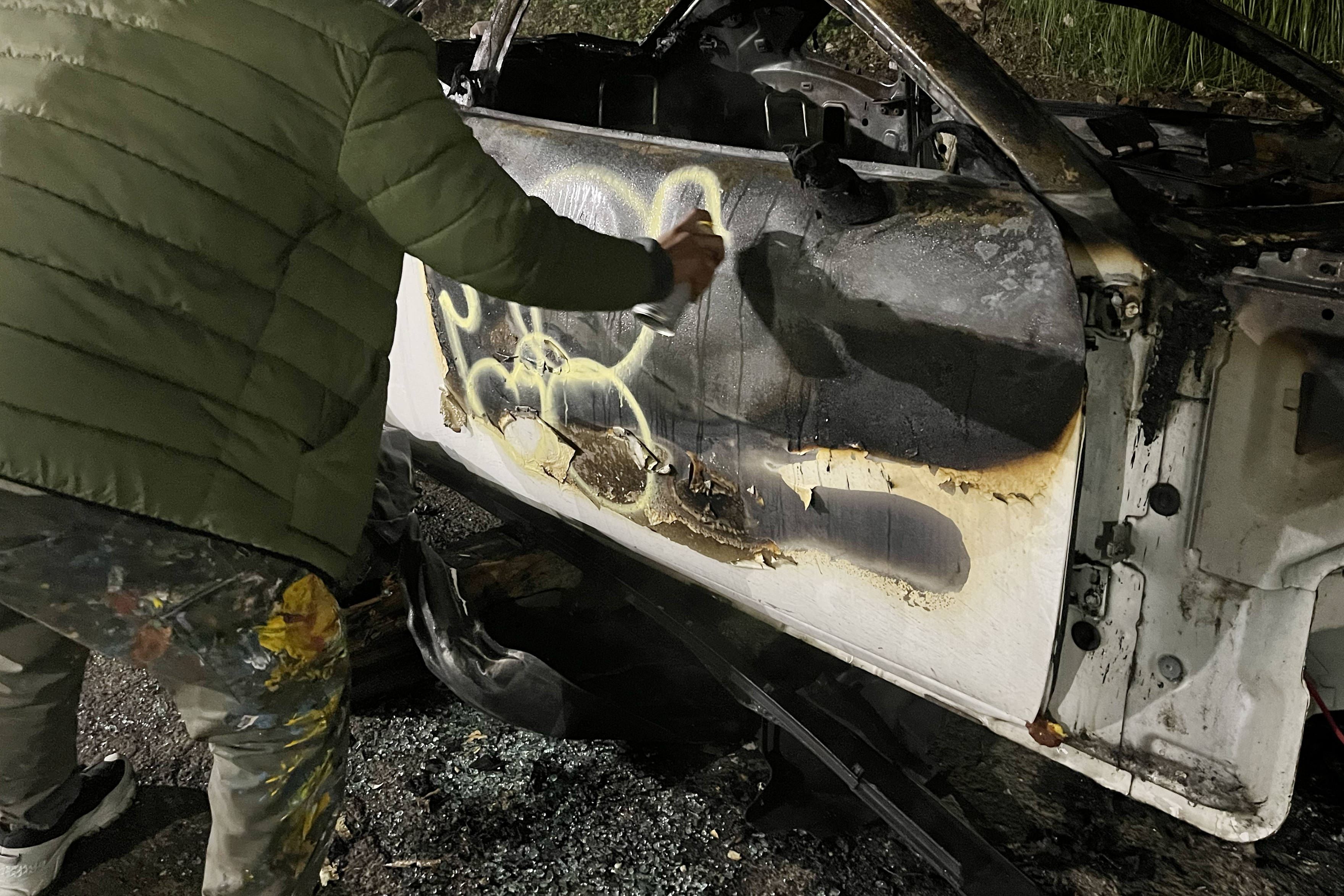 A person in a green jacket spray paints a yellow design on the charred side of a stripped, burnt vehicle. The ground is dark and the scene is lit by artificial light.