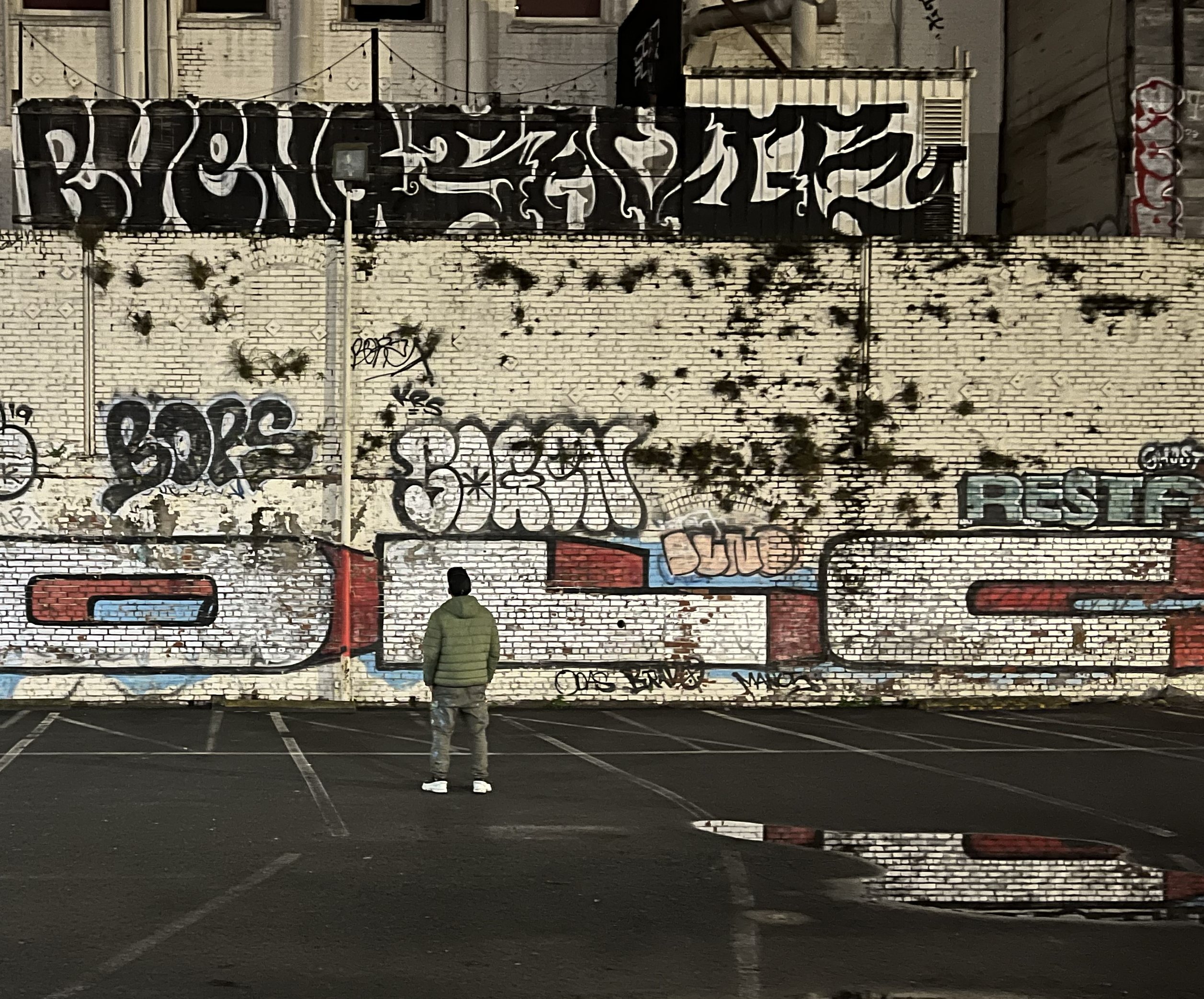 A person in a green jacket stands in an empty parking lot, facing a large graffiti-covered brick wall, with bold lettering and a reflective water puddle nearby.