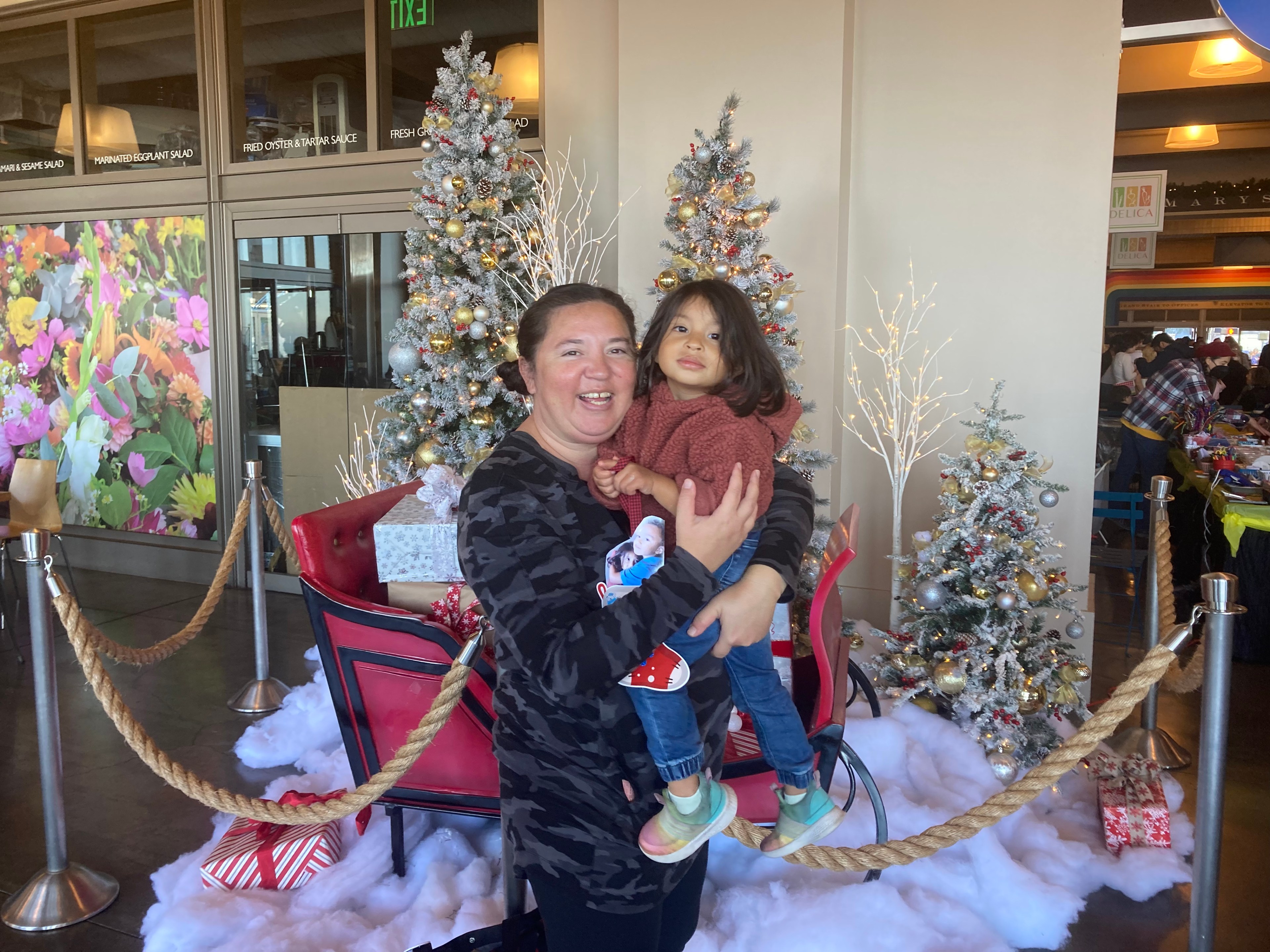 A smiling woman holds a child near decorated Christmas trees and a red sleigh. The background features festive decor and a colorful flower mural.