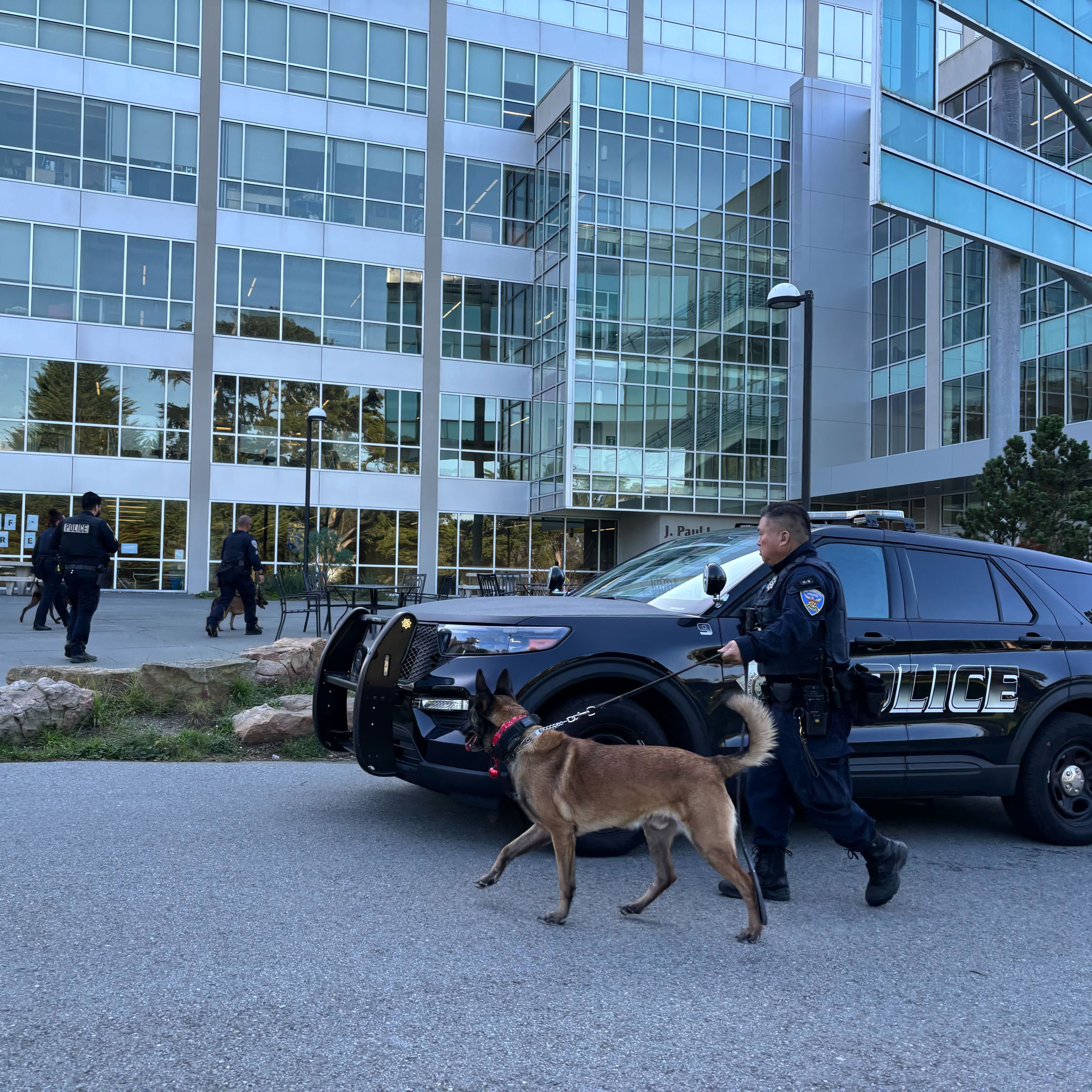A police officer walks a dog on a leash near a police SUV outside a modern glass building, with two officers in the background.