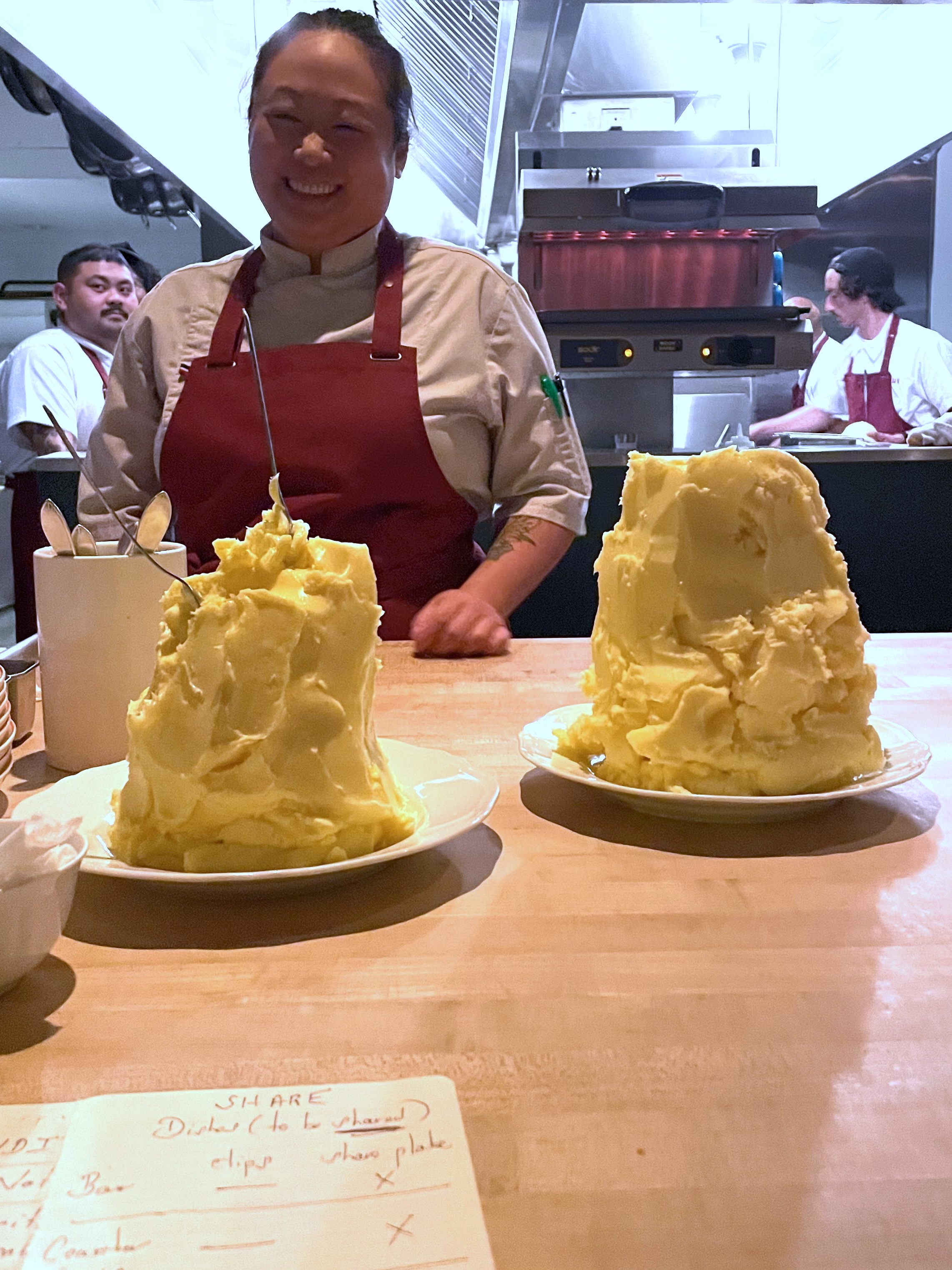 A smiling chef in a red apron stands behind two plates with large mounds of yellow butter. A kitchen with two other chefs is visible in the background.