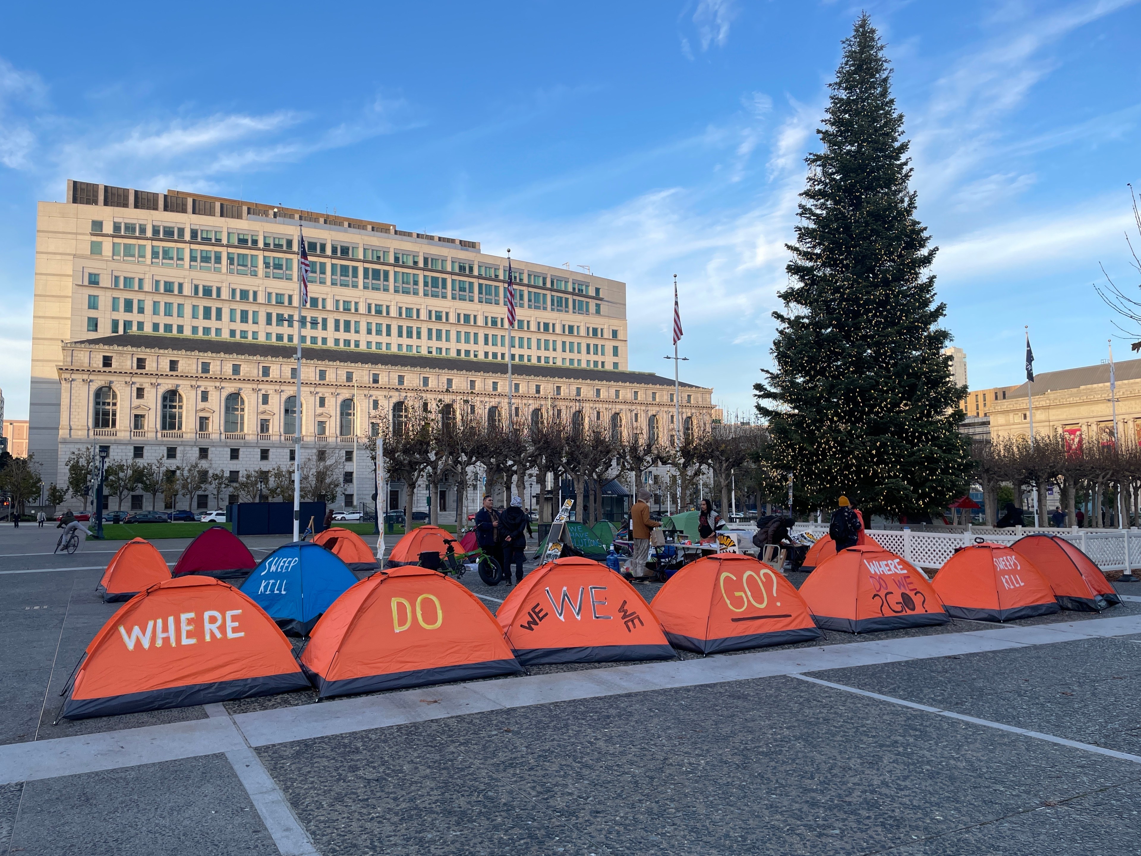 The image shows a row of orange tents with messages like &quot;WHERE DO WE GO?&quot; in front of a large, decorated Christmas tree and a grand building in the background.