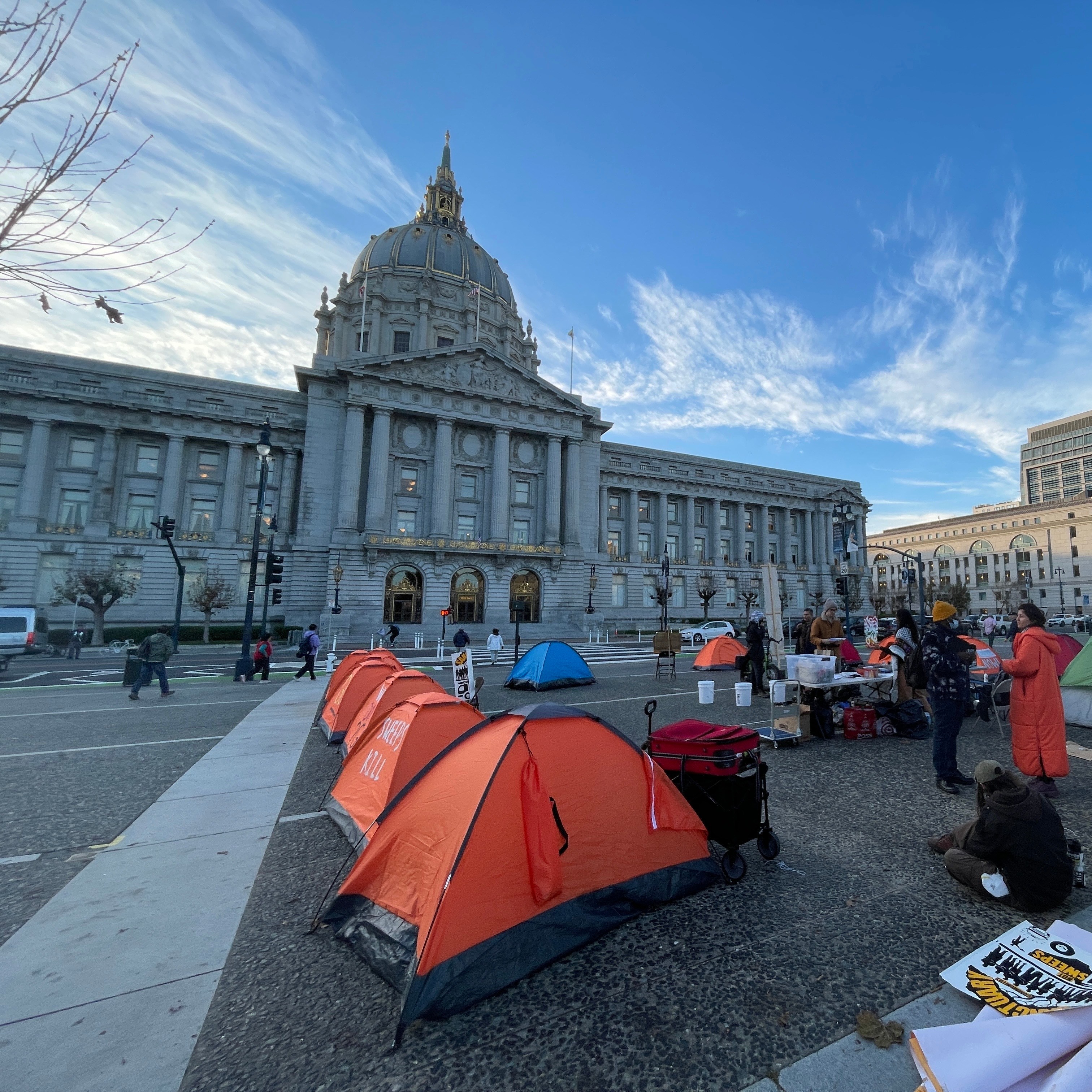 People gather around tents set up in front of a large, domed government building. The scene is lively, with a clear sky and modern buildings nearby.