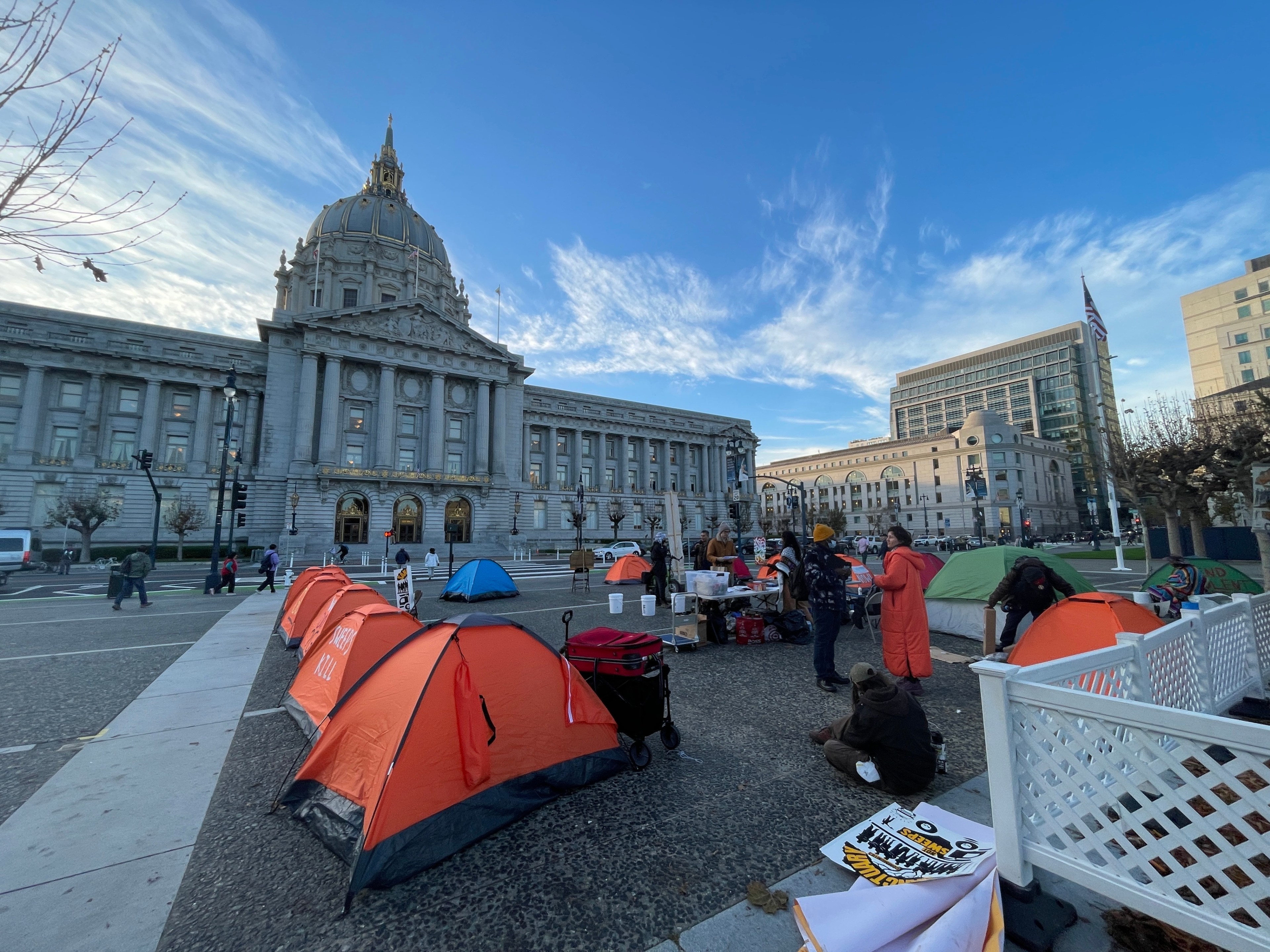 People gather around tents set up in front of a large, domed government building. The scene is lively, with a clear sky and modern buildings nearby.