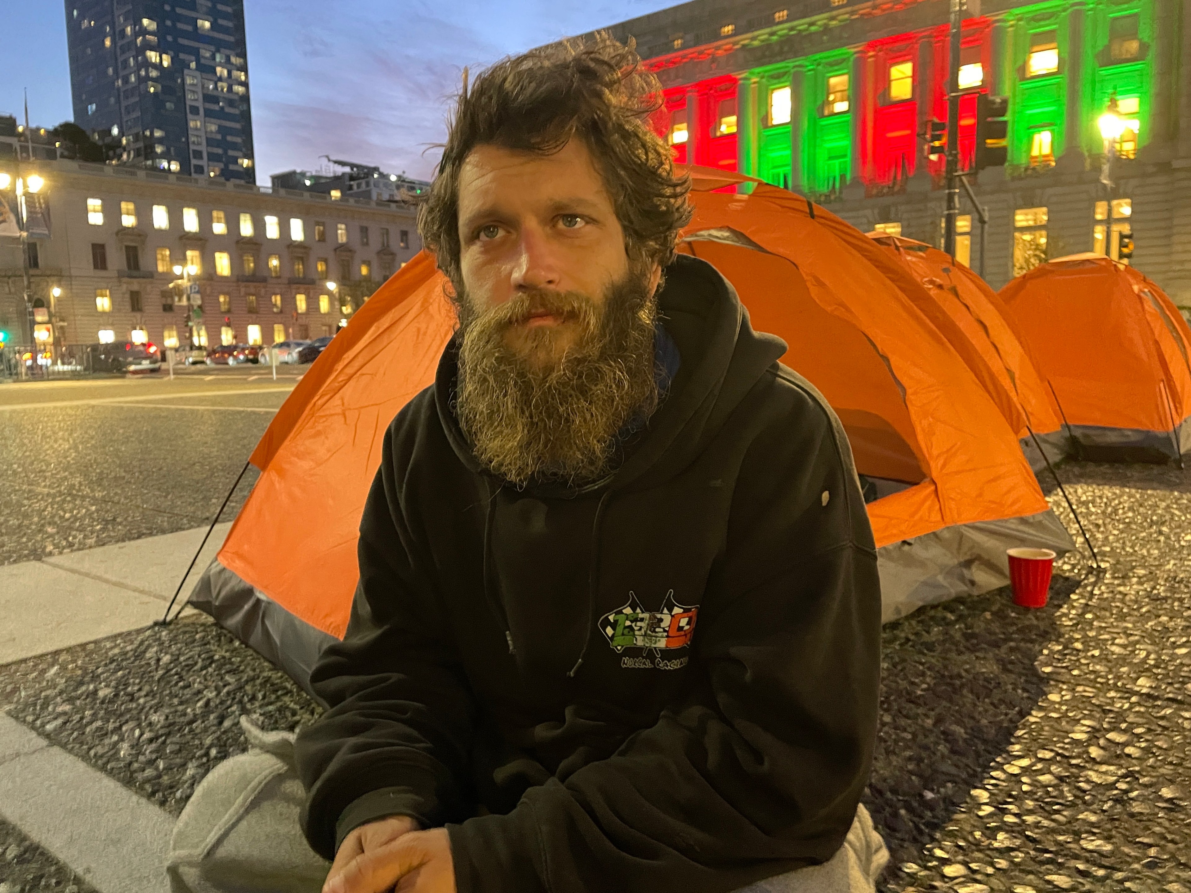 A bearded man in a black hoodie sits outside an orange tent in a lit city square at dusk. Buildings with illuminated windows and colorful lights are in the background.