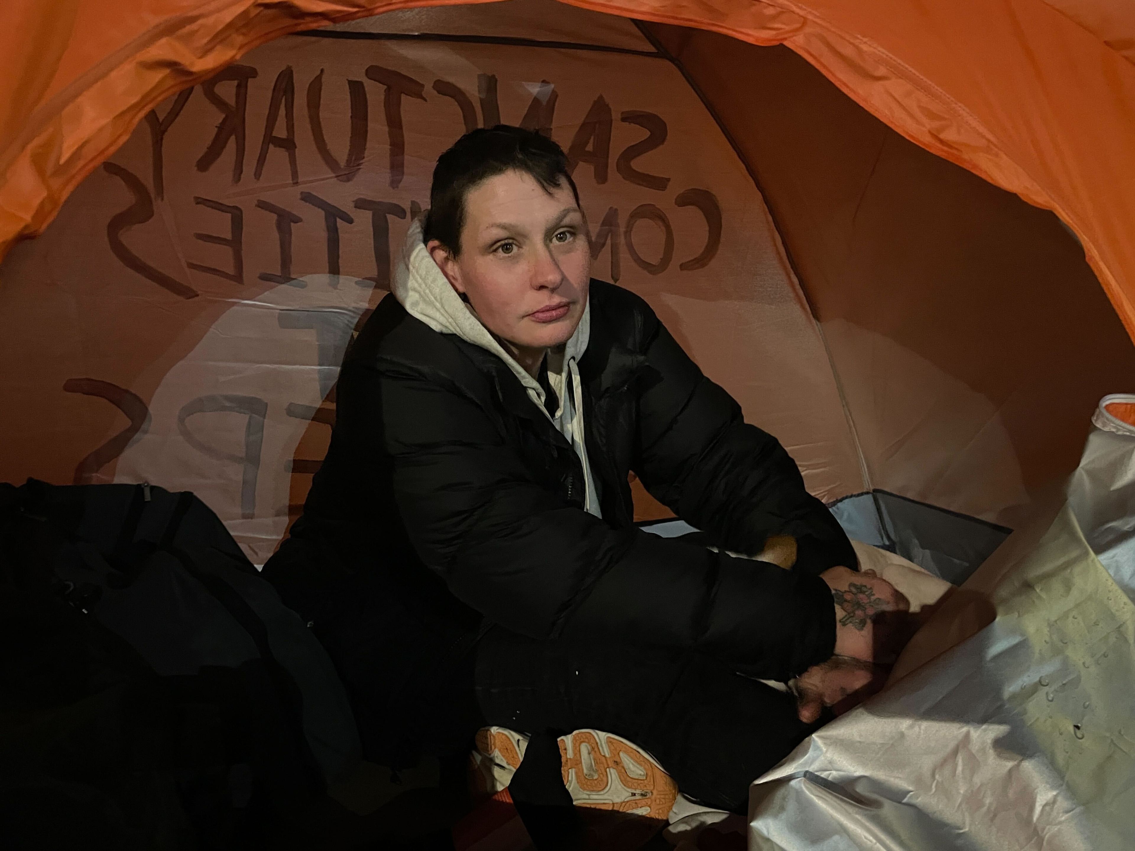 A person sits inside an orange tent, wearing a black jacket with a white hood. They appear thoughtful, and the tent has writing on the interior.